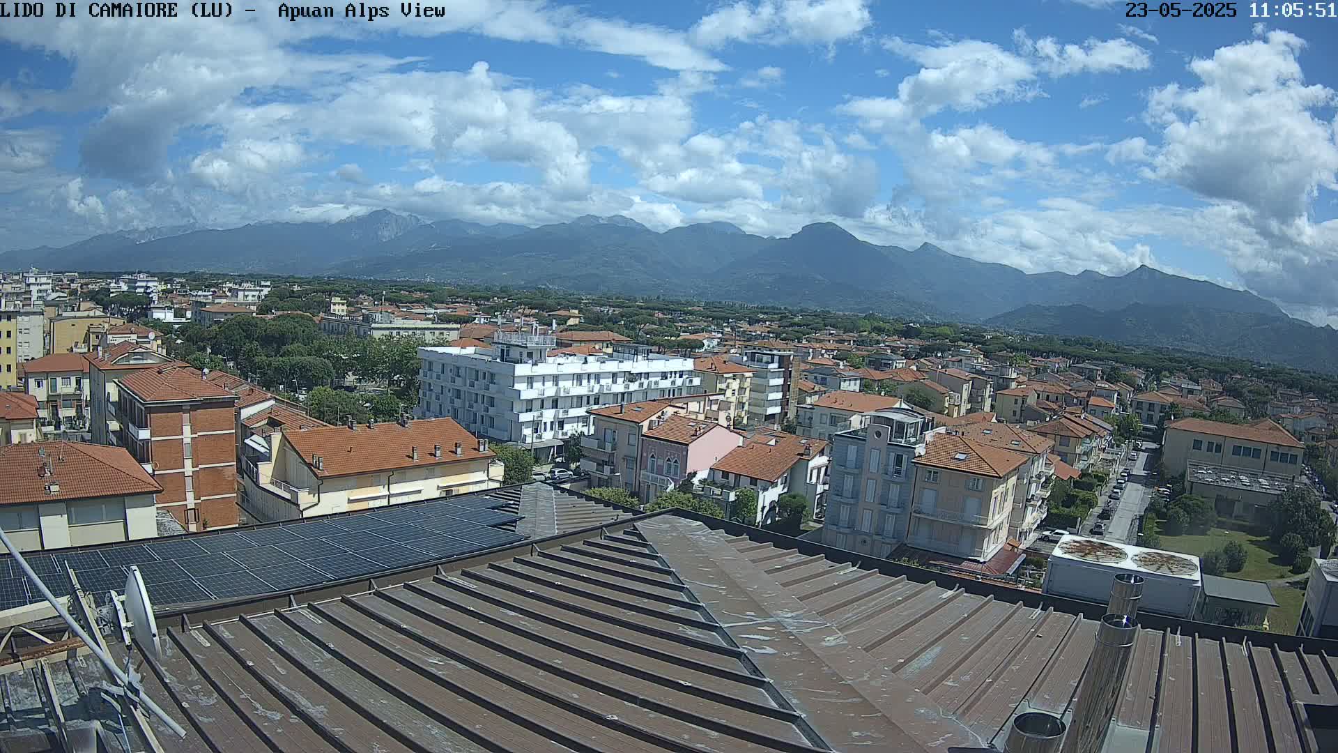A partly cloudy day reveals a cityscape nestled beneath a mountain range, viewed from a rooftop with solar panels.