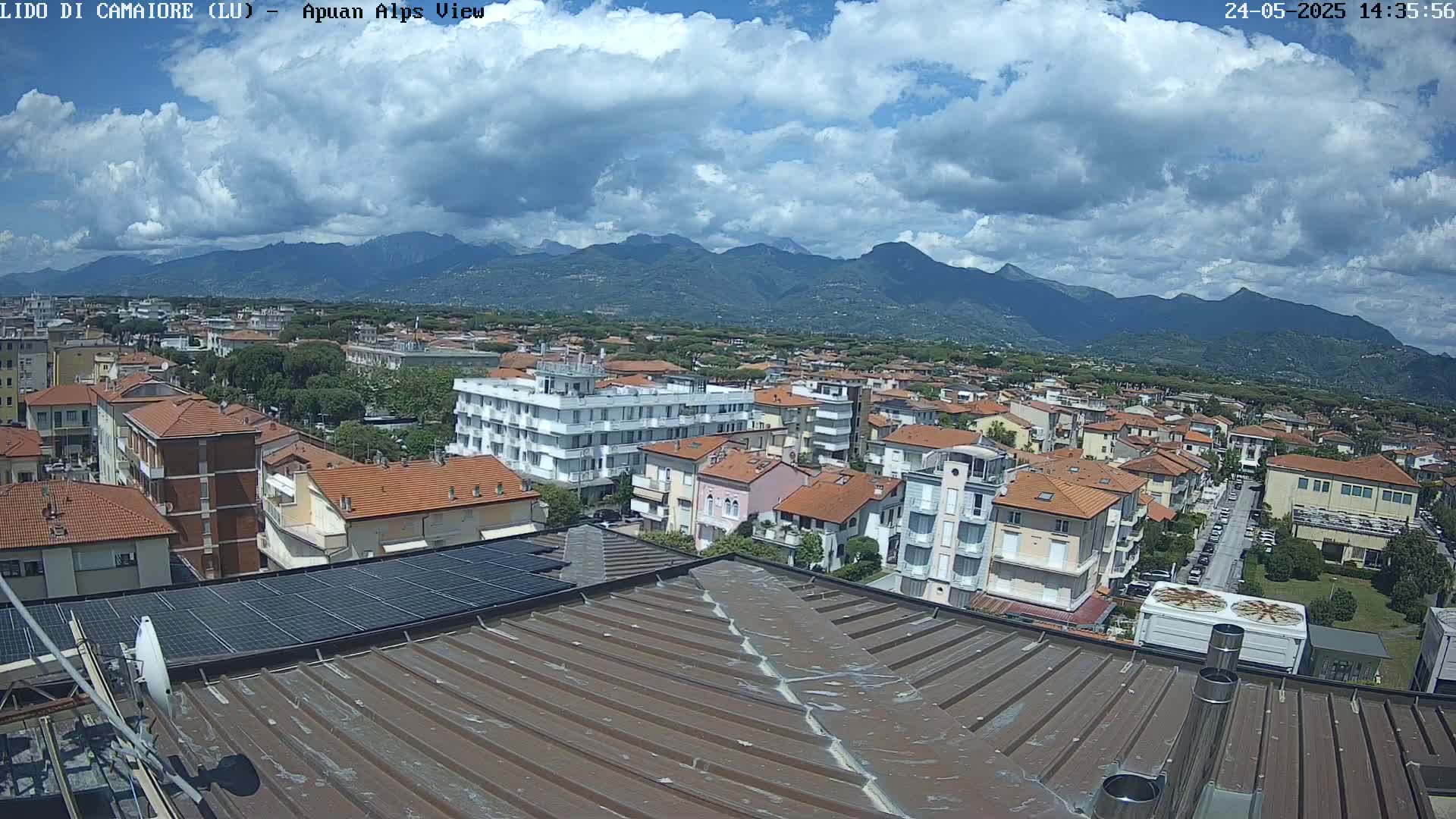 A partly cloudy day reveals a sprawling town nestled at the foot of a mountain range, viewed from a rooftop with solar panels and satellite dish.