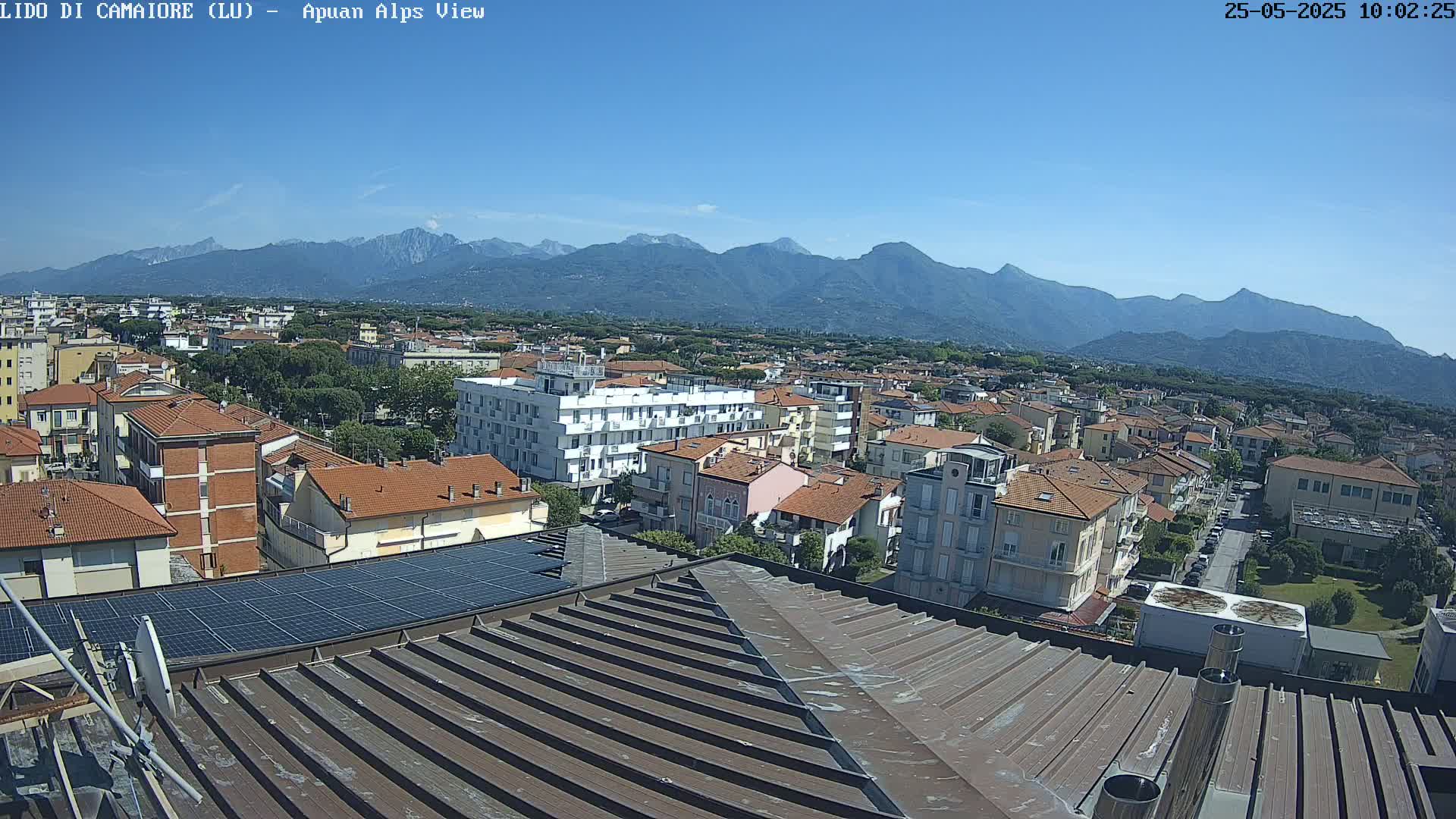 A view from a rooftop shows a town stretching towards a mountain range under a clear blue sky.