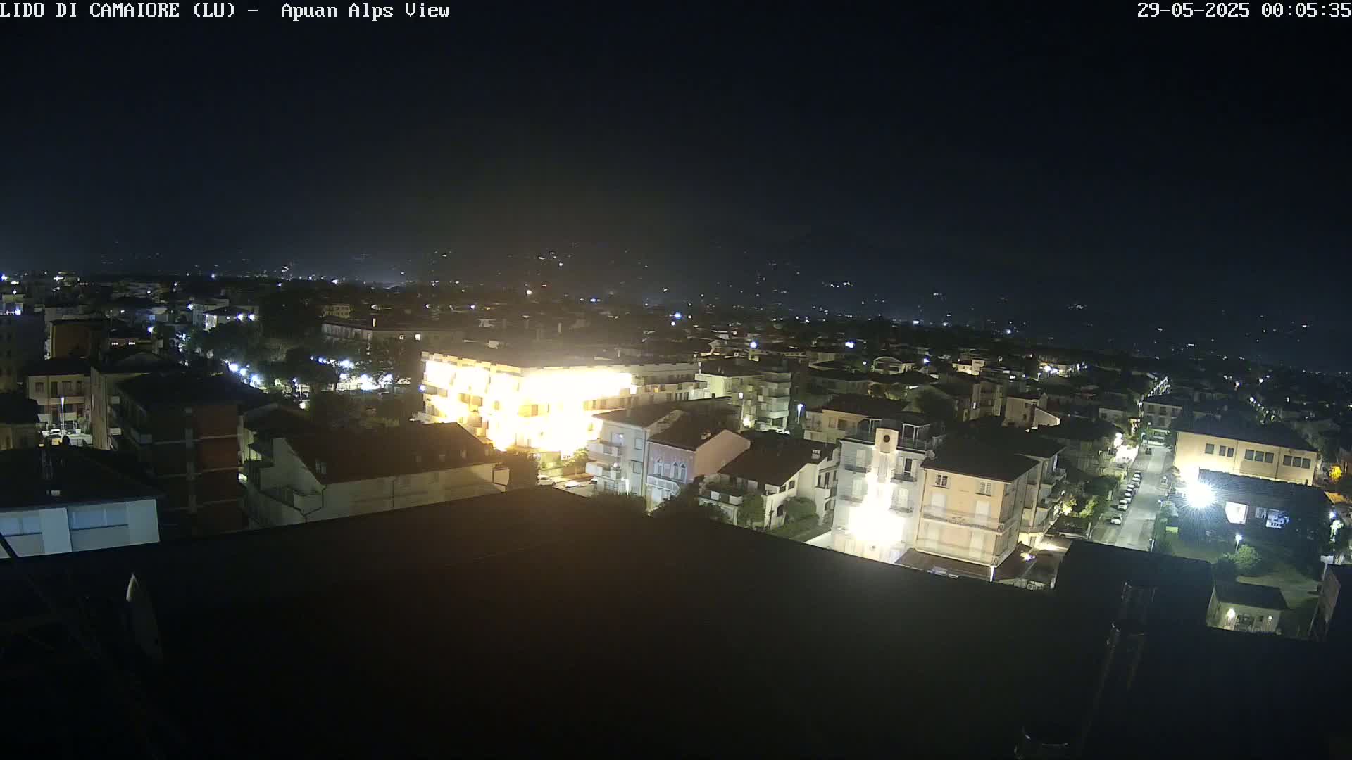 A nighttime, high-angle view of a town illuminated by streetlights, with a dark sky and distant hills faintly visible in the background.