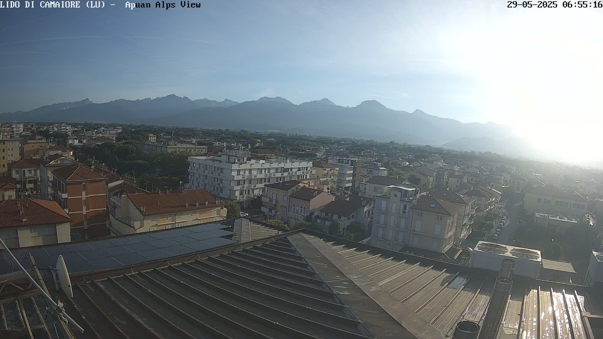 A hazy sunrise illuminates a cityscape nestled at the base of a mountain range, viewed from a rooftop with solar panels.