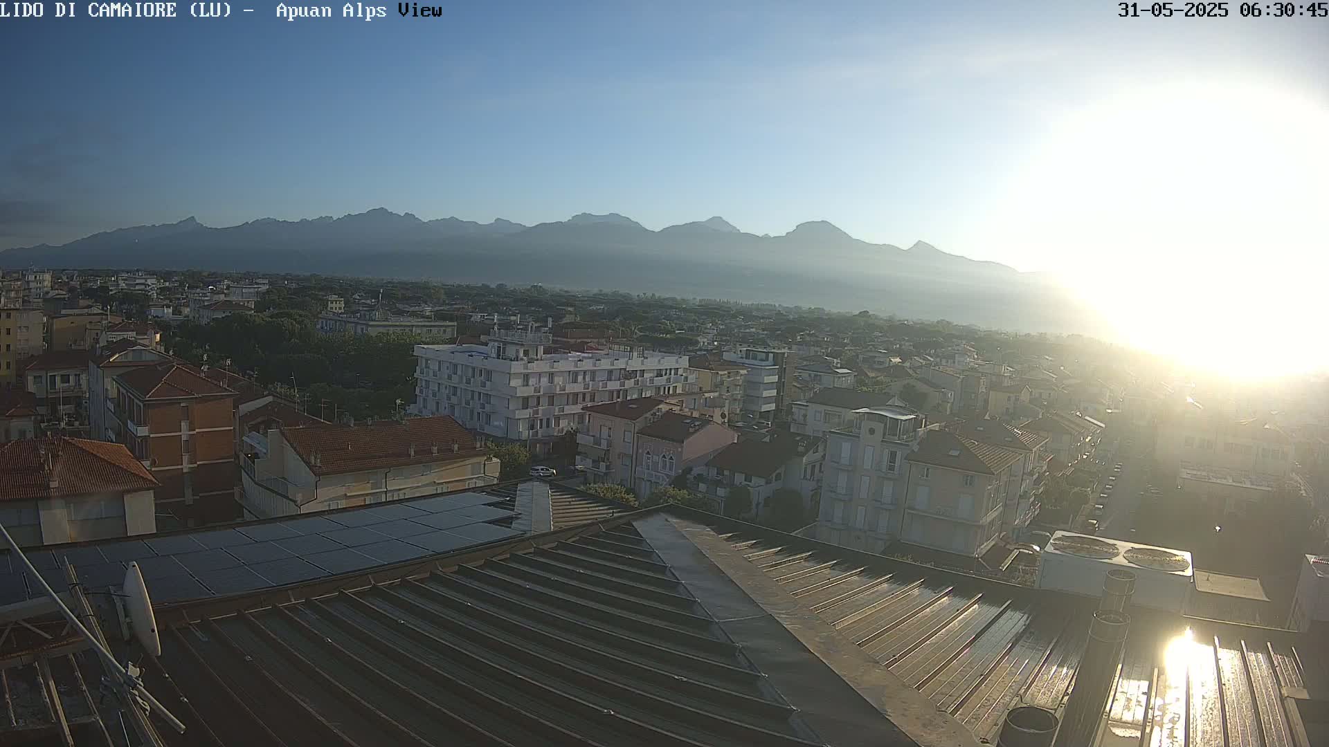 A hazy sunrise illuminates a town nestled beneath a mountain range, viewed from a rooftop with solar panels.