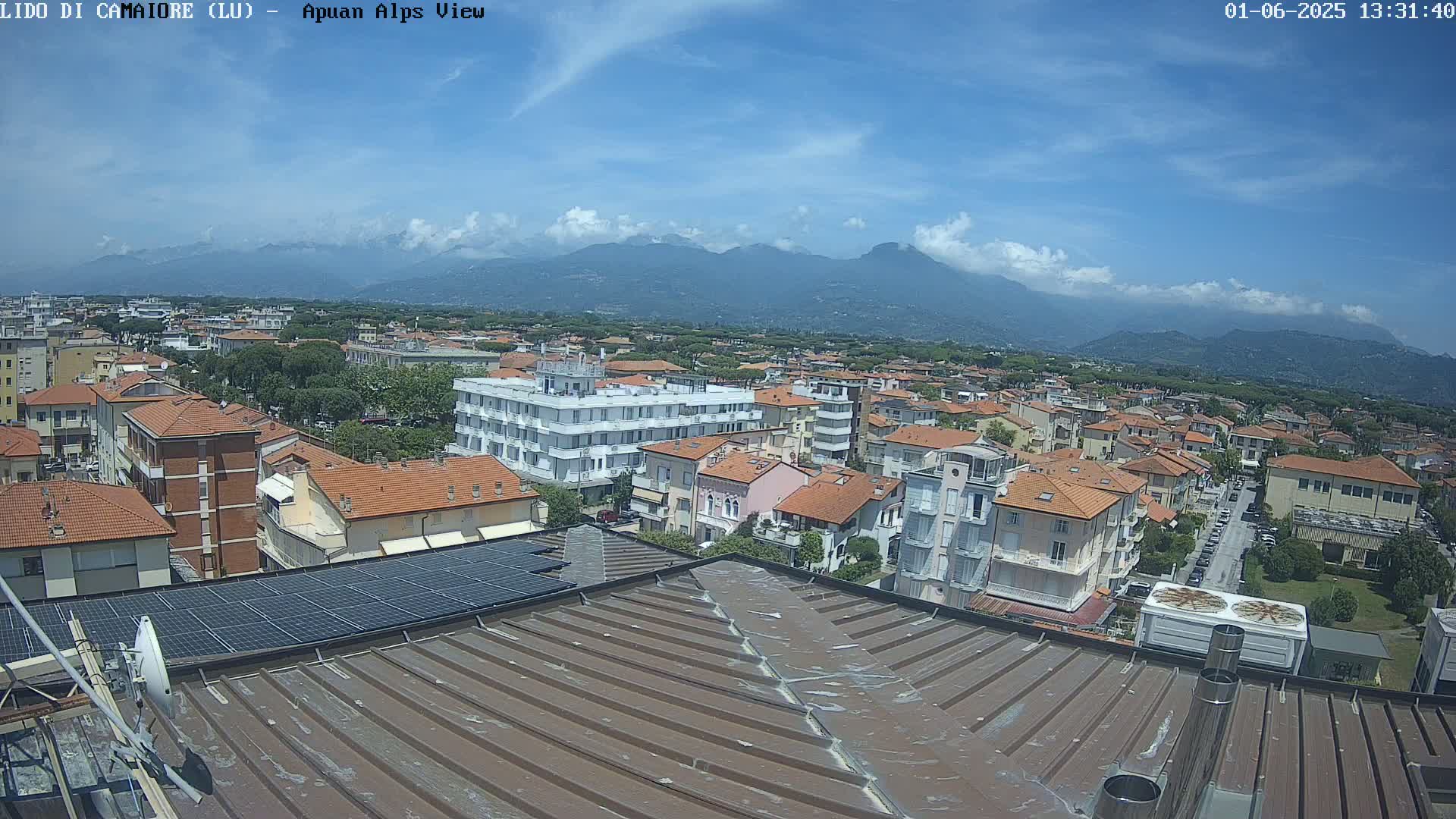 A rooftop view of a town nestled against a mountain range under a mostly sunny sky with some scattered clouds.
