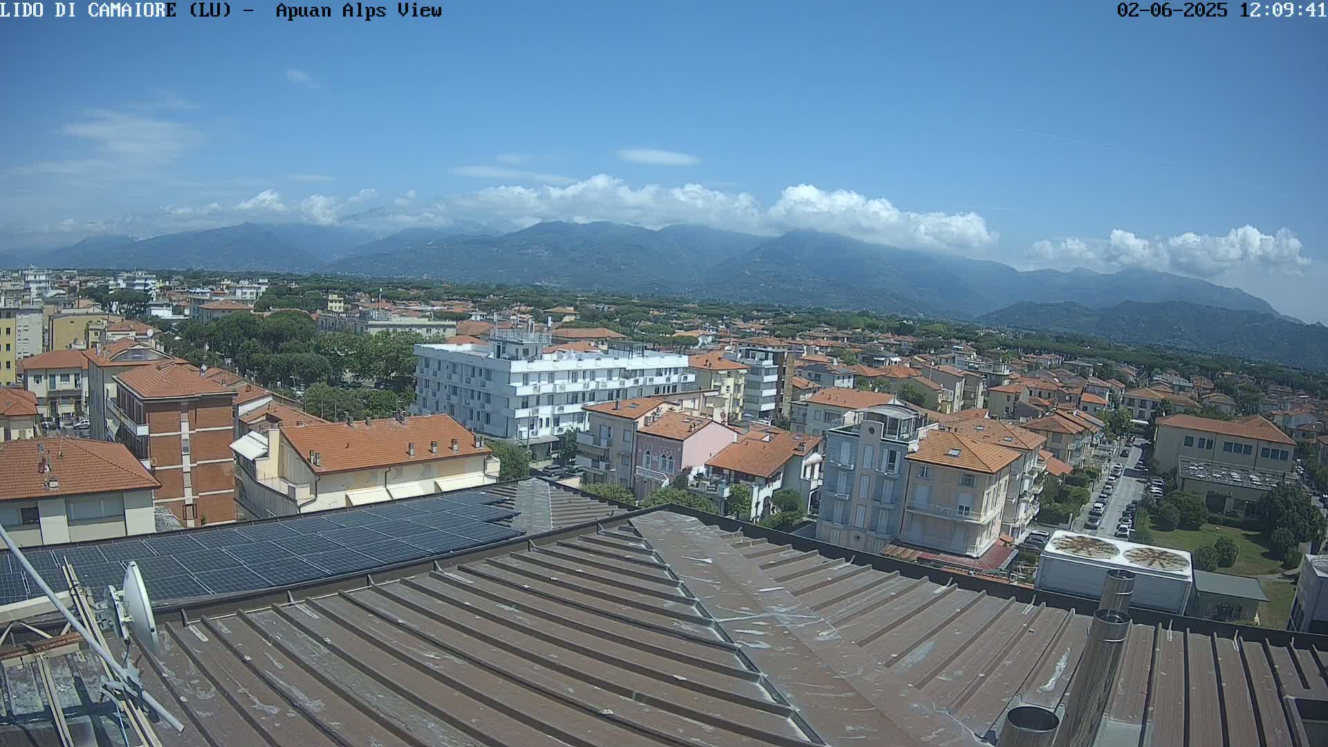 A mostly sunny day reveals a rooftop view of a town nestled against a mountain range with some puffy clouds in the sky.