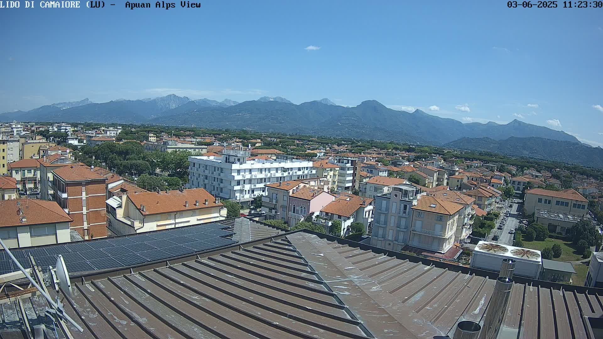 A sunny day reveals a view of a town nestled at the foot of a mountain range, seen from a rooftop with solar panels.