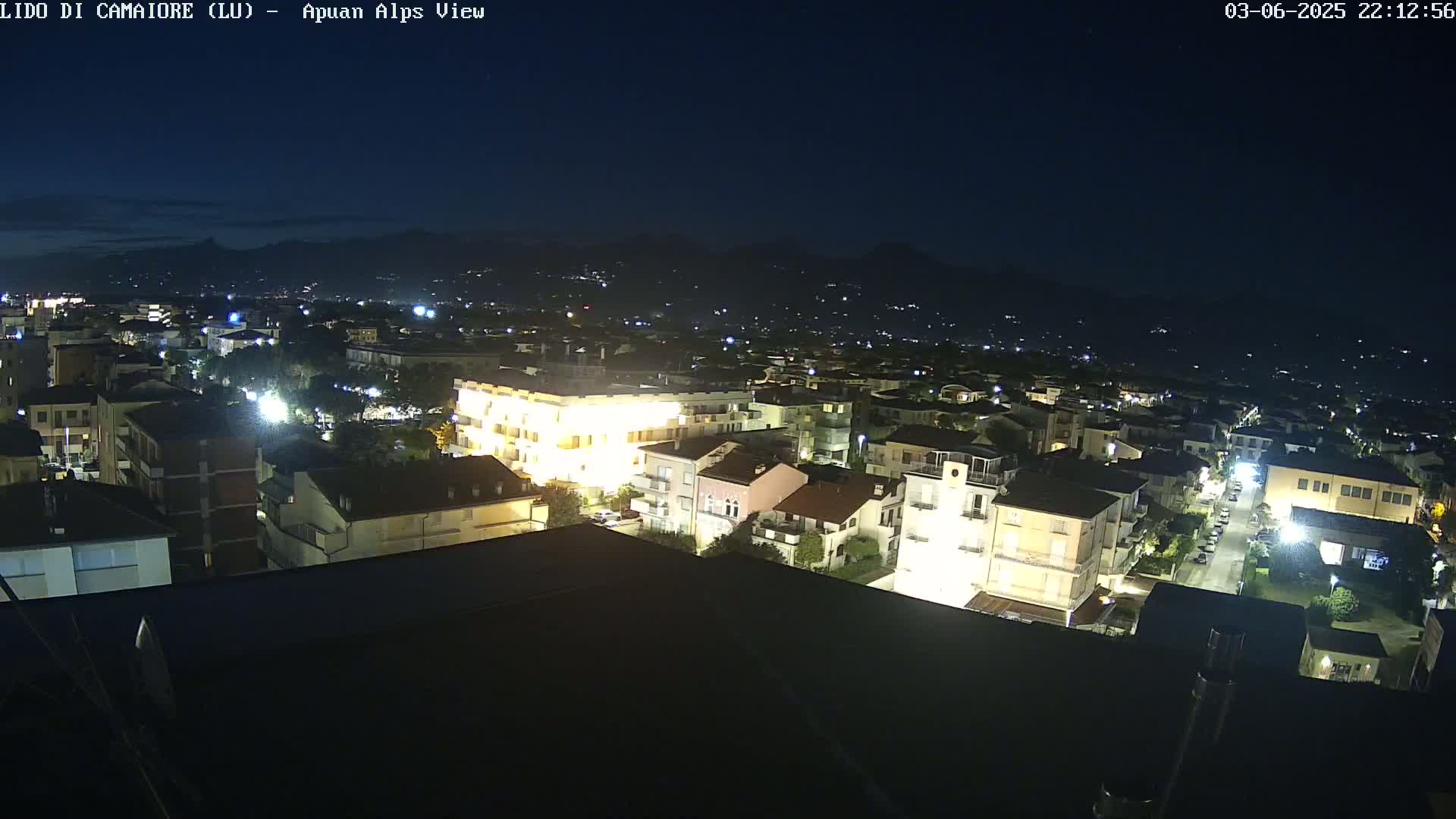 A nighttime, aerial view shows a town illuminated with streetlights, with dark mountains visible in the distance under a clear, dark sky.