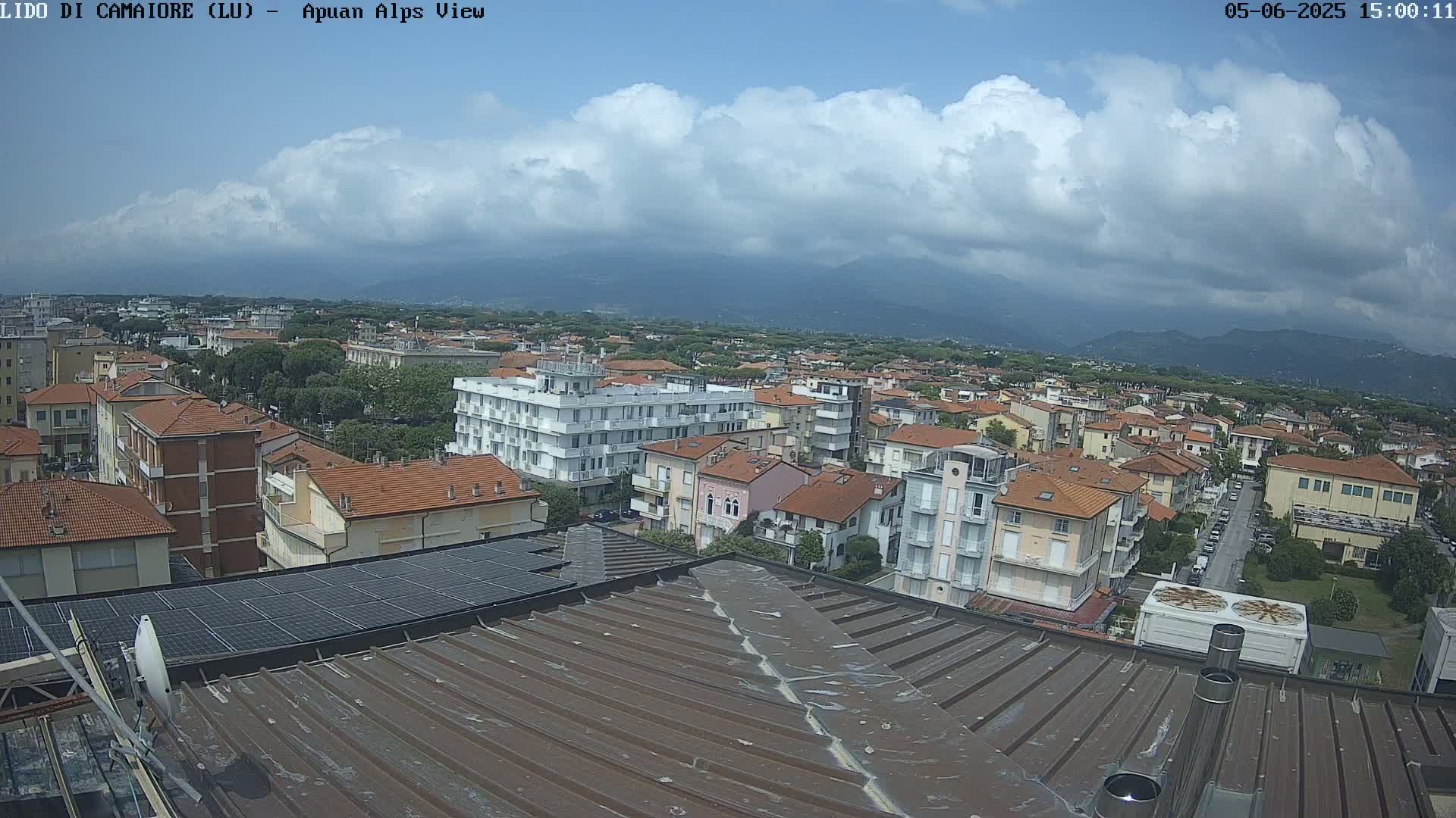 A partly cloudy day overlooks a coastal town with numerous buildings, a rooftop solar panel array, and distant mountains.