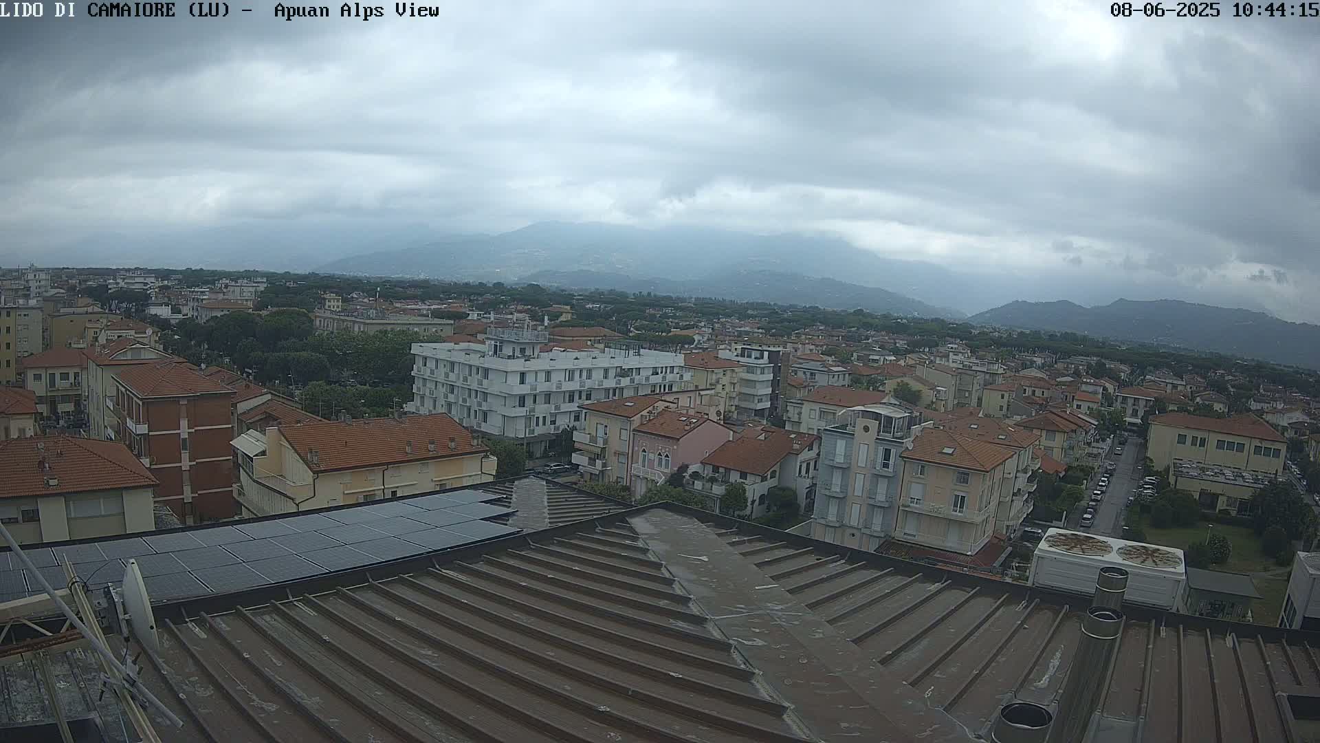 A rooftop view of a town under a cloudy sky, showing many buildings and distant mountains.
