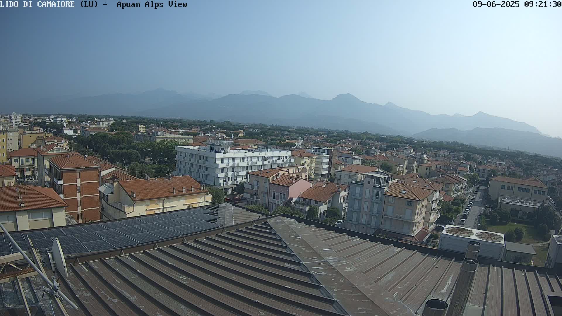 A hazy view of a town, with a mountain range in the background, is seen from a rooftop with solar panels.