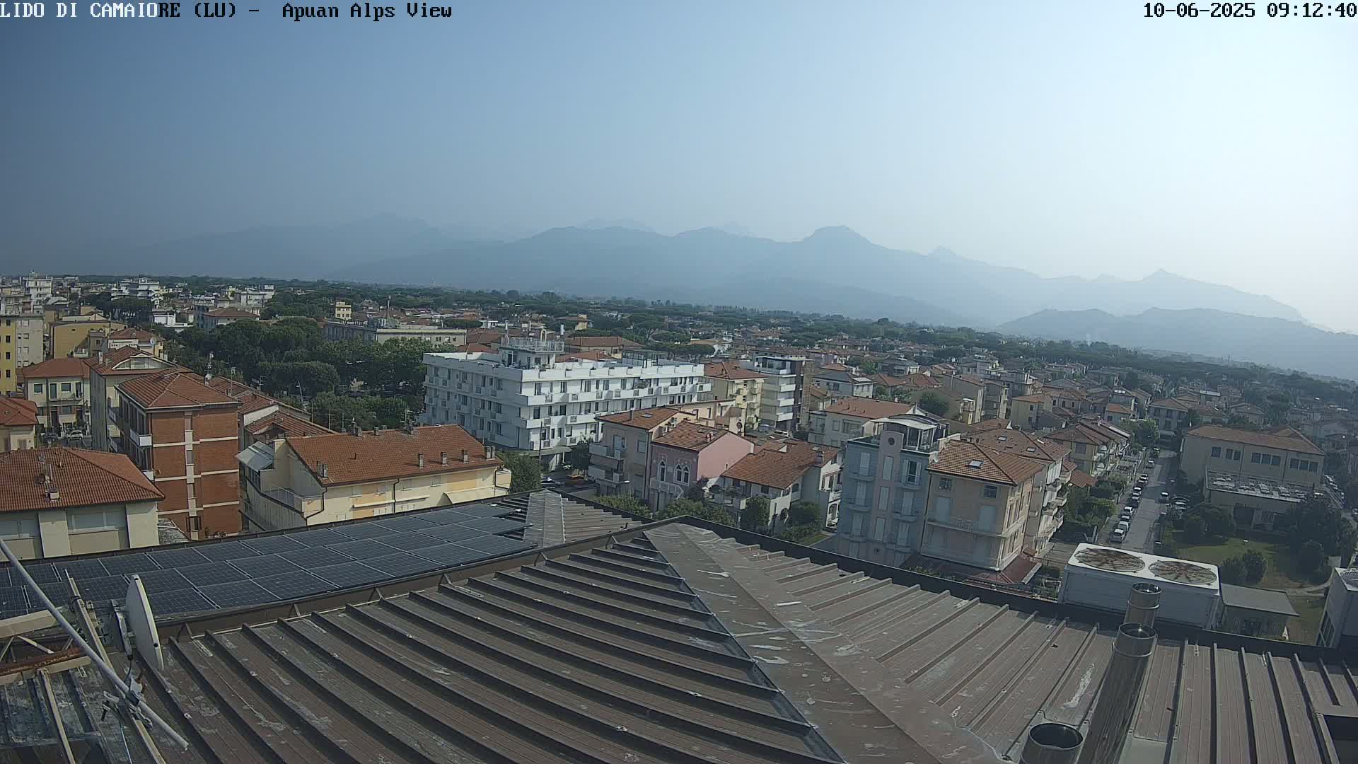 A hazy view of a town with red-tiled roofs and taller buildings, nestled against a range of mountains under a clear sky.