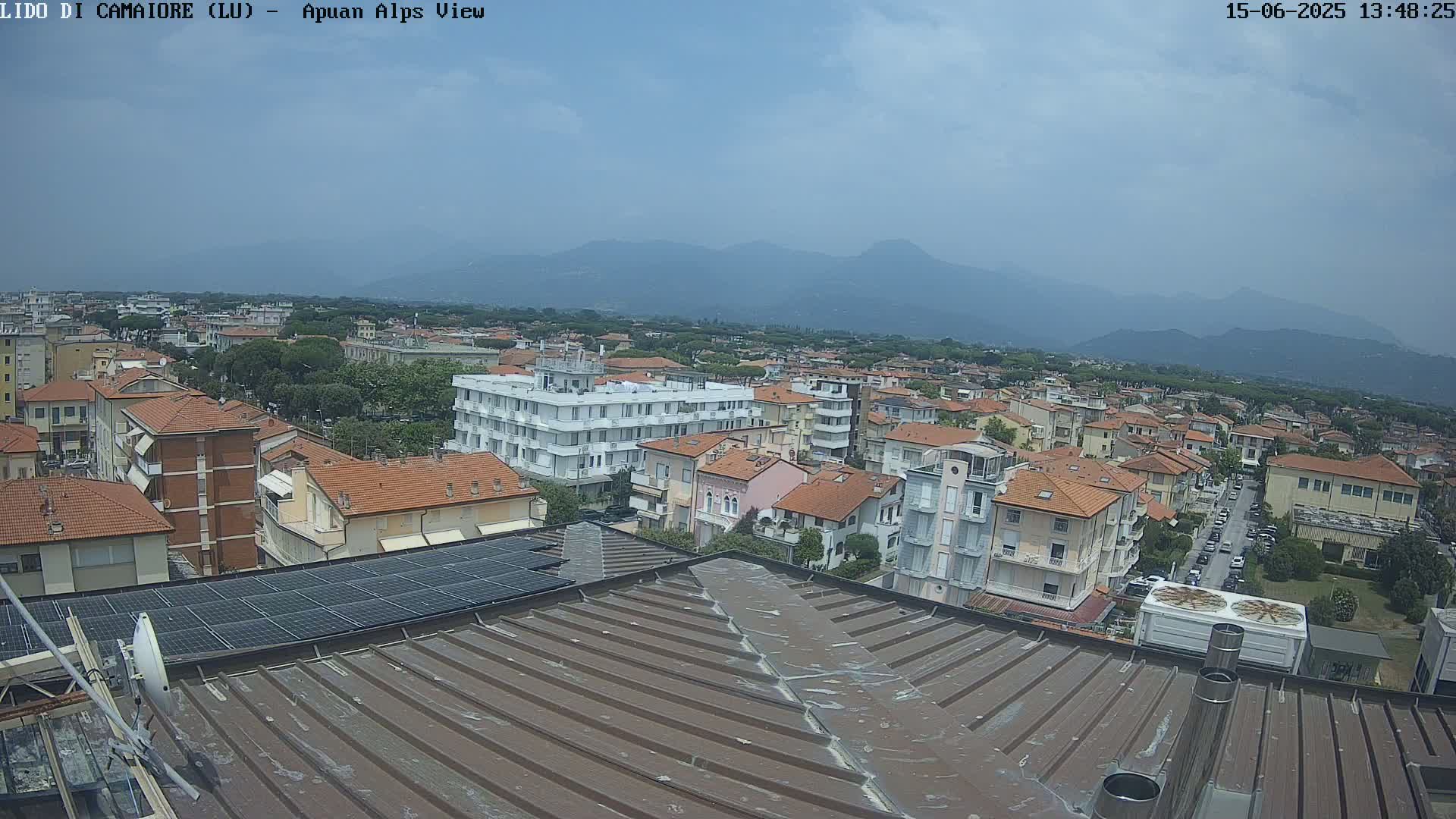 A slightly hazy view of a town with red-tiled roofs, partially visible mountains in the background, and a rooftop with solar panels in the foreground under a partly cloudy sky.
