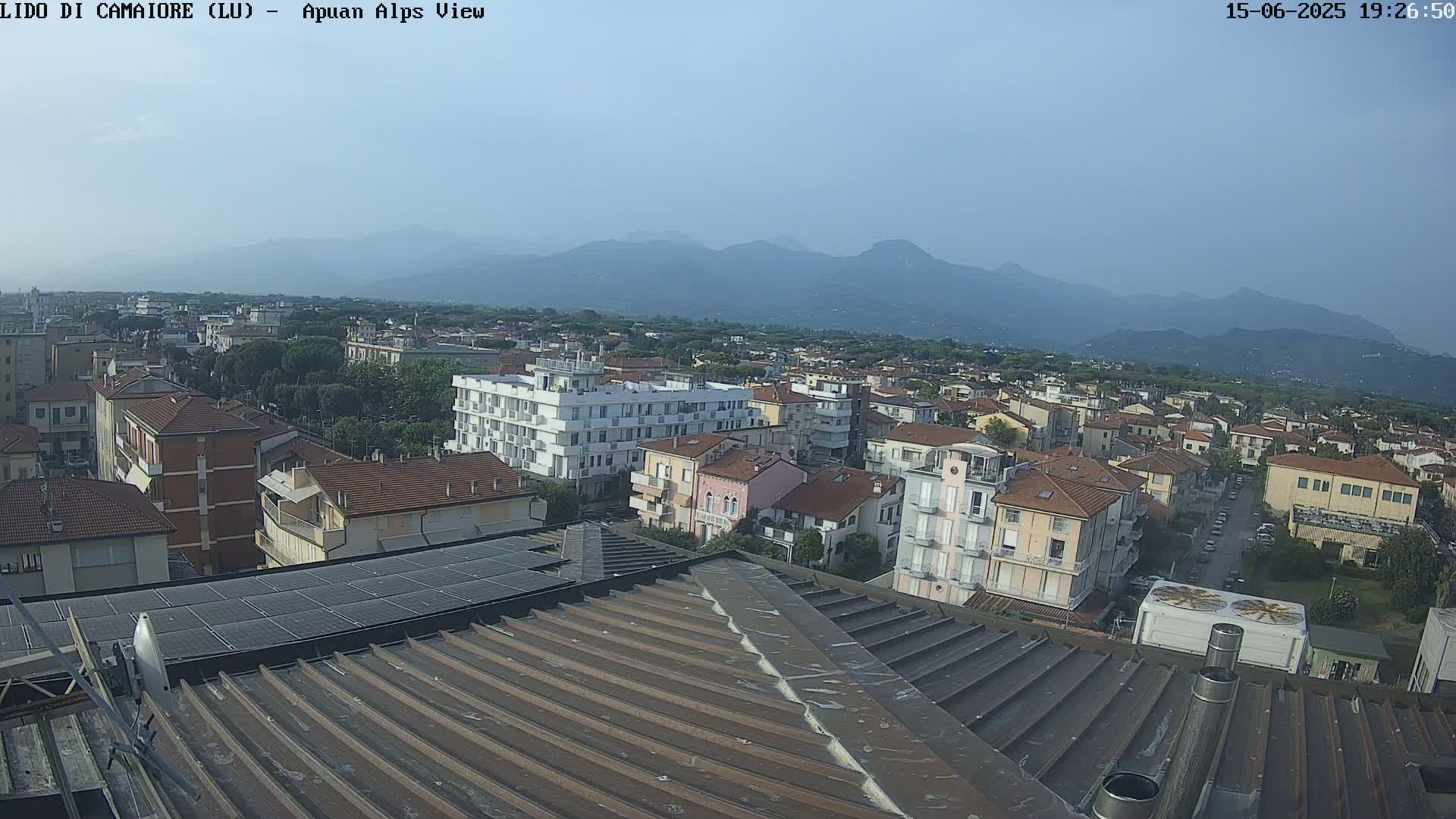 A hazy, sunny day overlooks a town with numerous buildings, a solar panel array, and distant mountains.