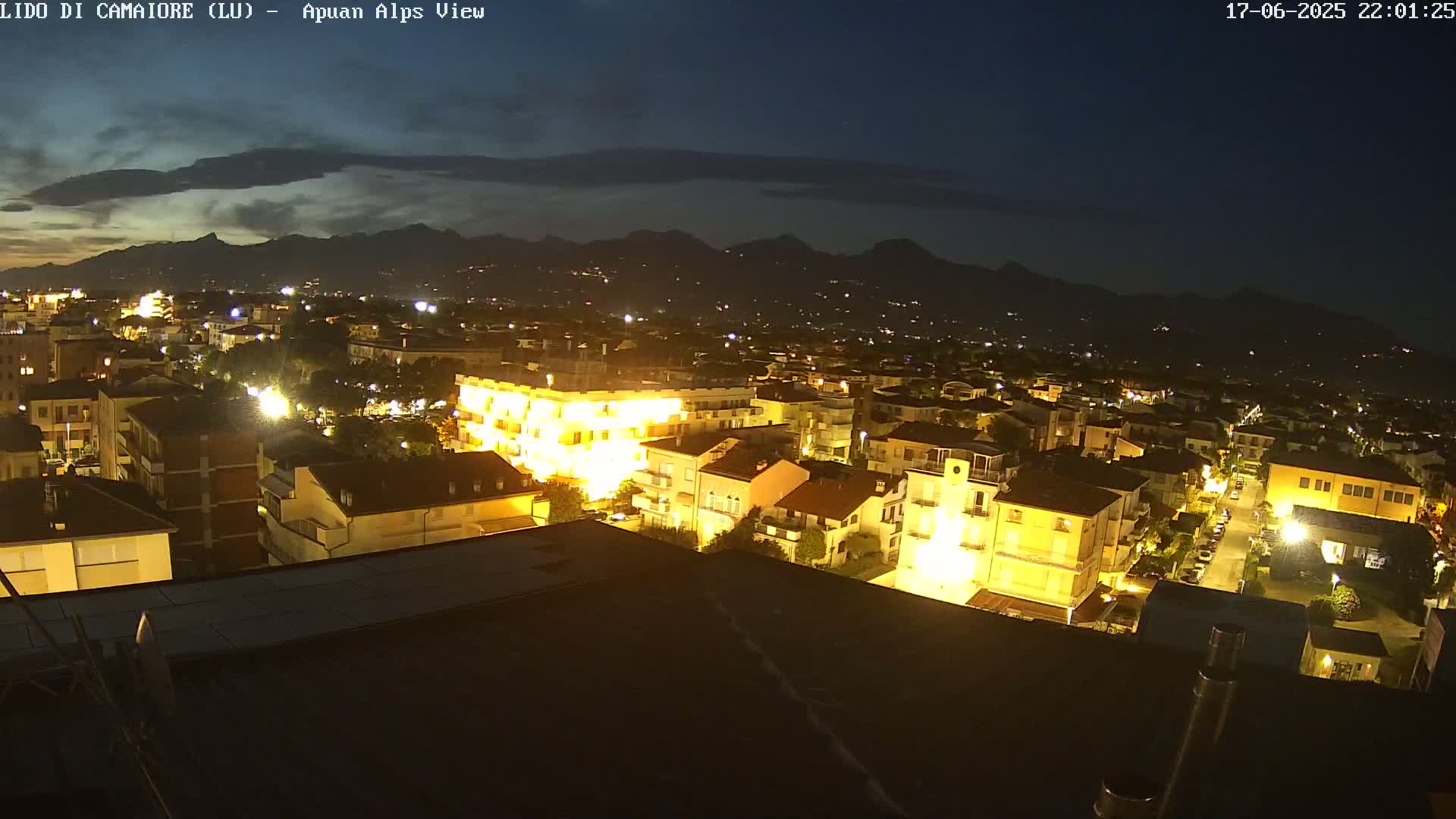 A nighttime, high-angle view of a town illuminated by streetlights, with mountains in the background under a partly cloudy sky.