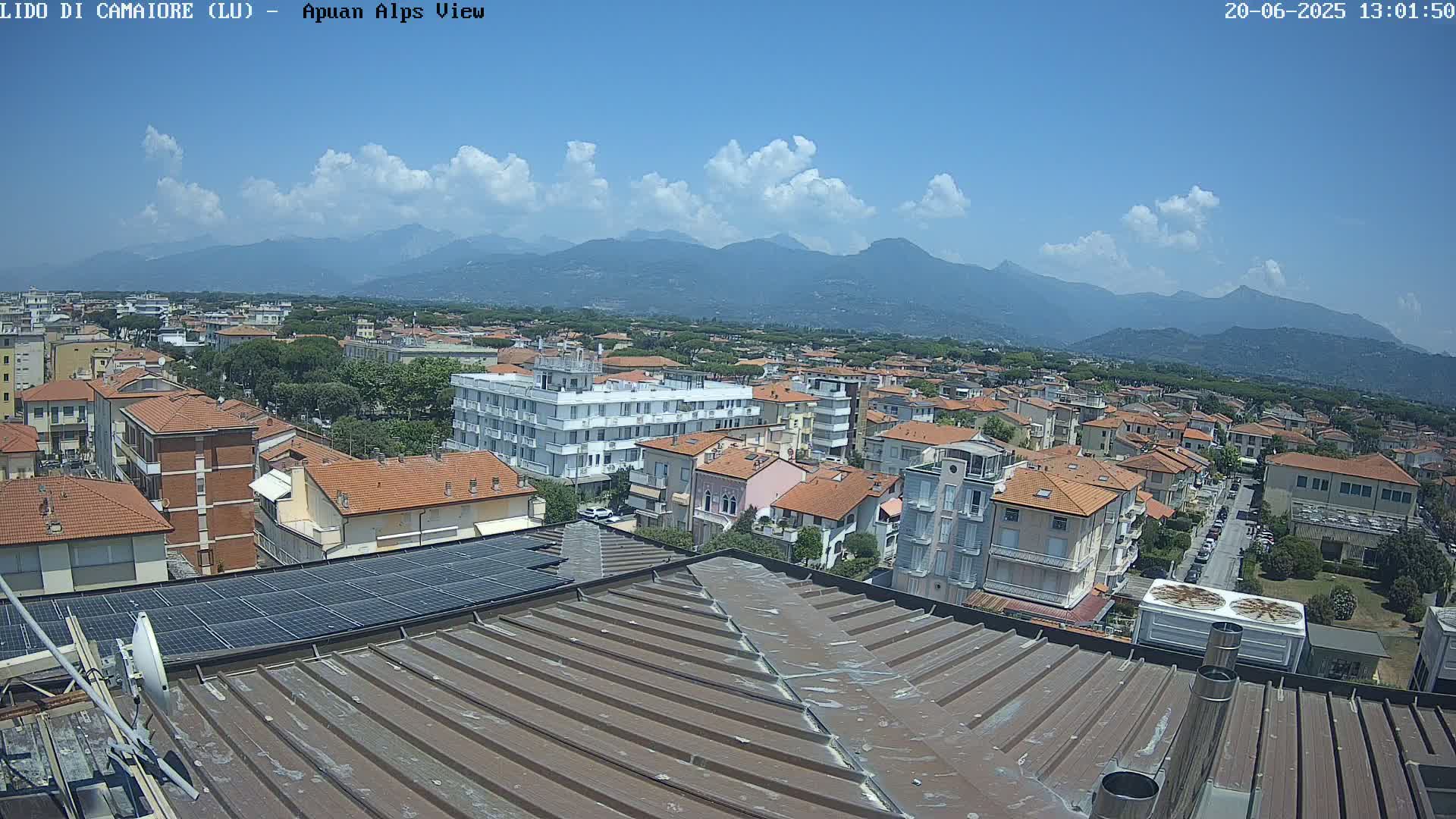 A rooftop view shows a town with red-tiled buildings under a partly cloudy sky, with mountains in the distance.