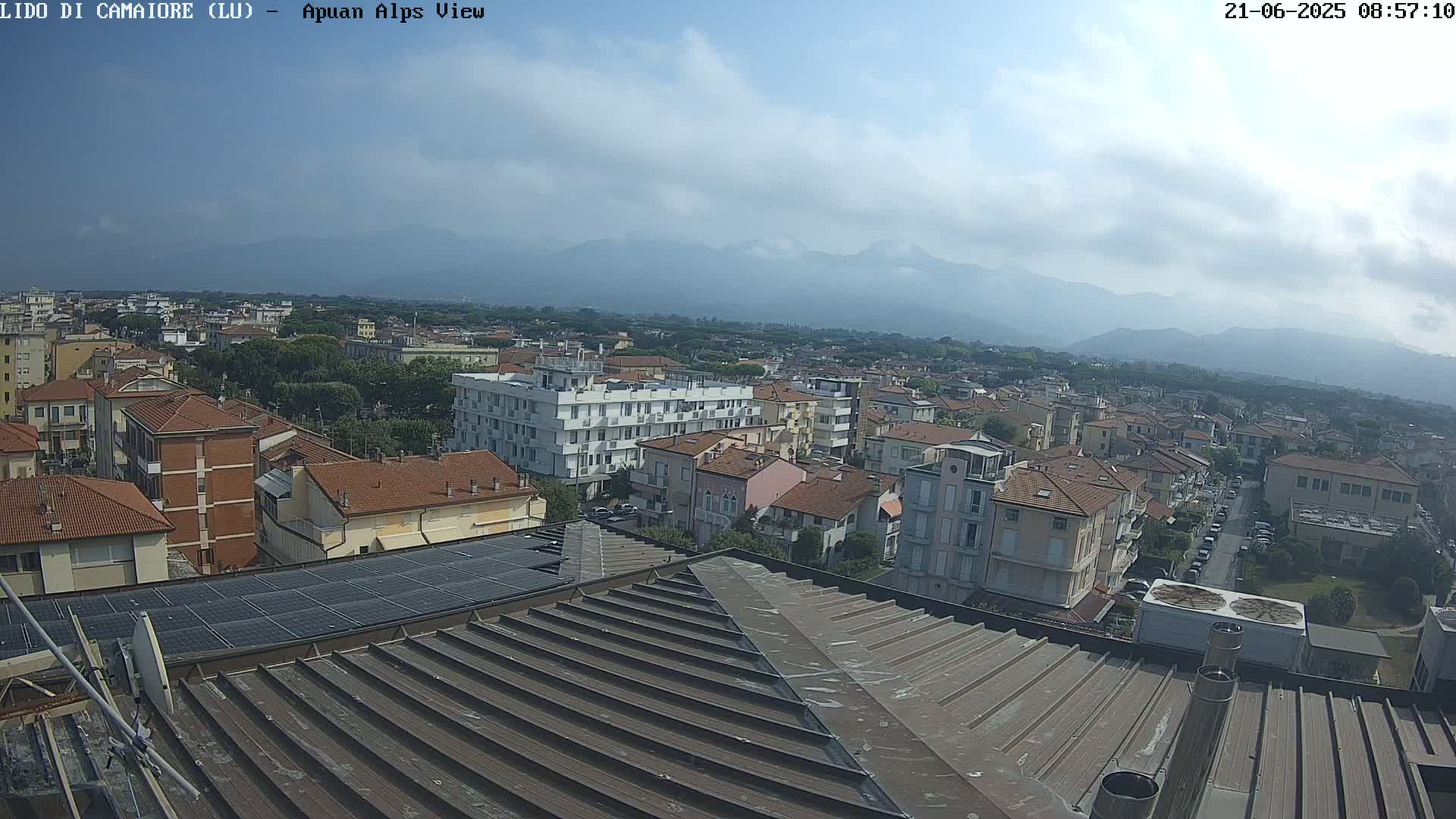 A view of a town in the Apuan Alps, under a partly cloudy sky, shows many low-rise buildings and a rooftop with solar panels.