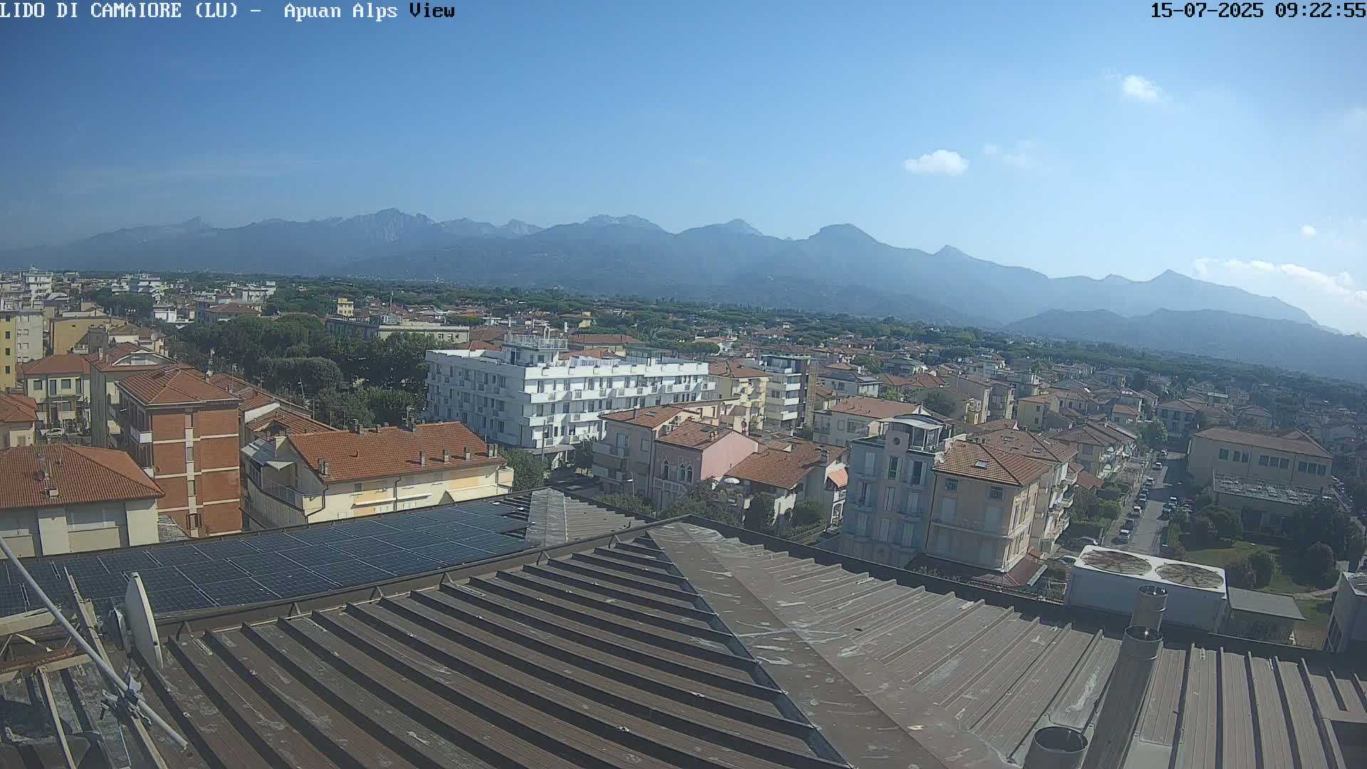 A sunny day reveals a cityscape nestled beneath a mountain range, with a view from a rooftop showing buildings and solar panels.