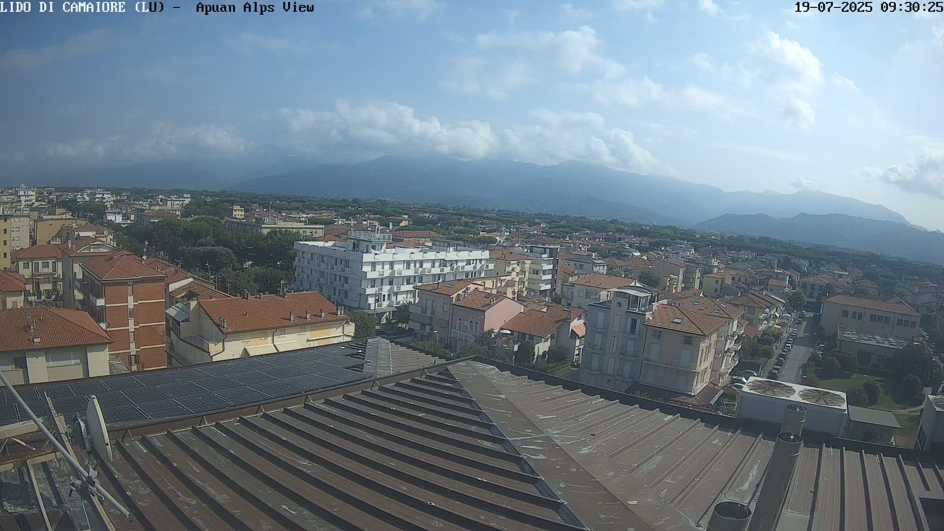 A partly cloudy day reveals a view of a town nestled against a mountain range, seen from a rooftop with solar panels.