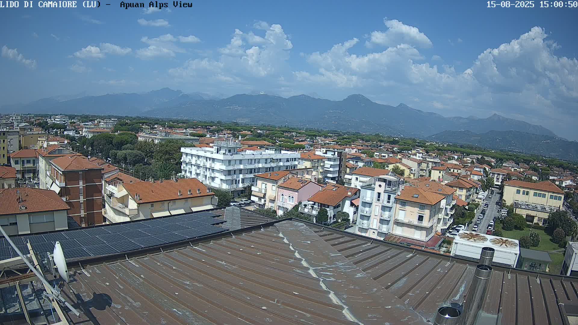 A mostly sunny day reveals a view of a town nestled below a mountain range, seen from a rooftop with solar panels.