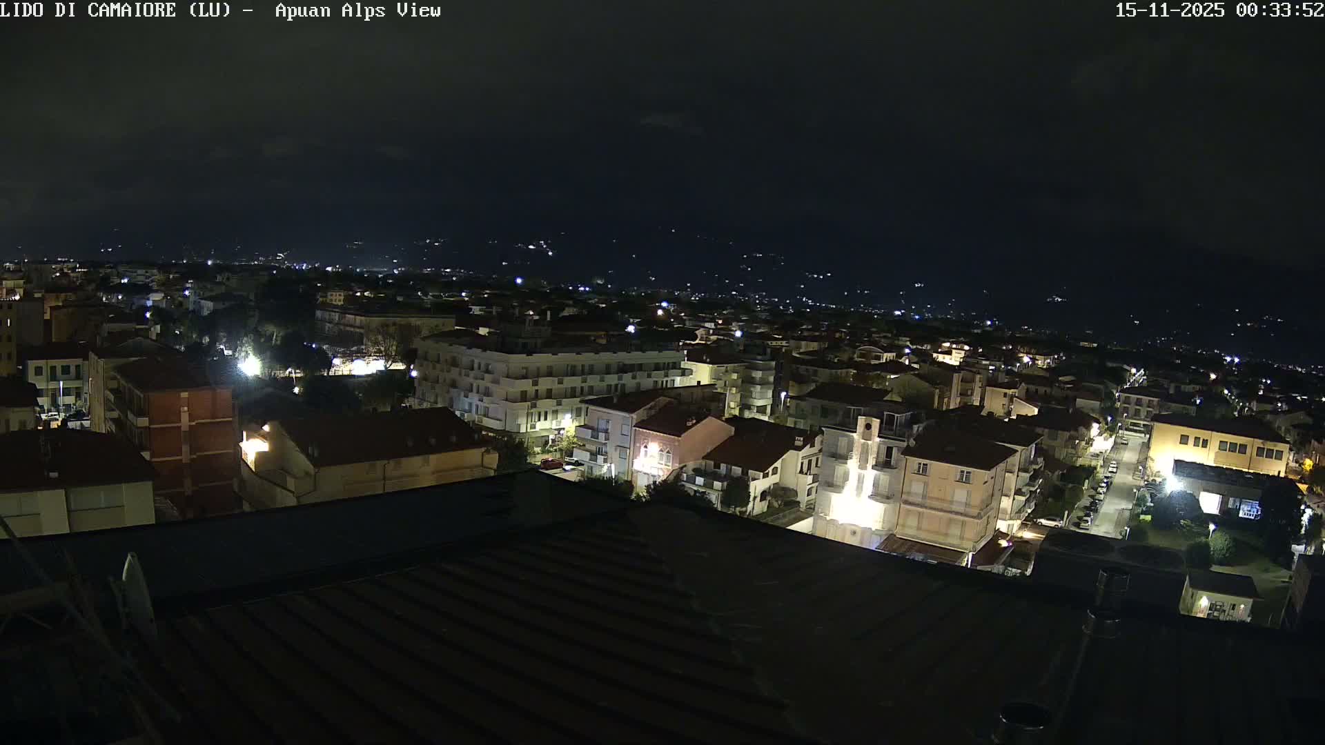 A clear night reveals a brightly lit town or city sprawling across the landscape, with numerous buildings and streetlights illuminating the scene, and distant lights visible against a dark sky with some cloud cover.