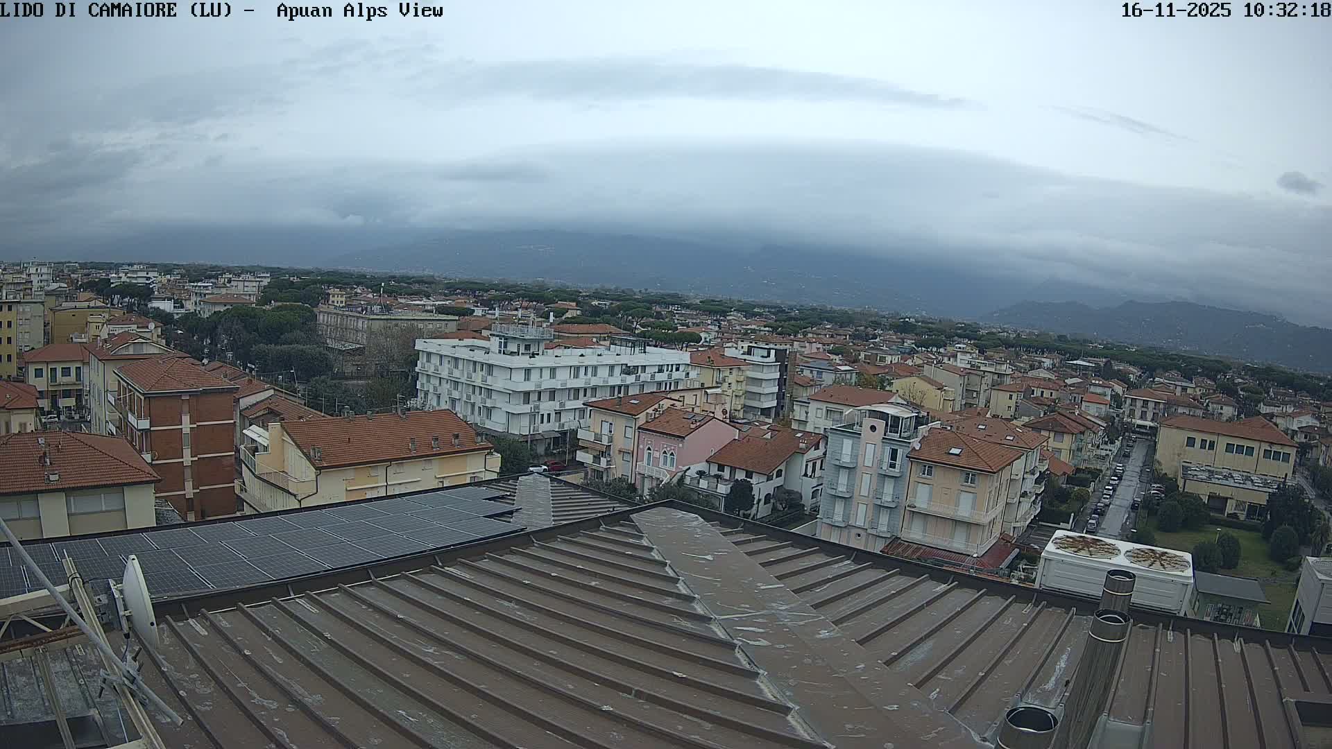A high-angle view reveals a densely built town with varied architecture and solar panels, extending towards distant, hazy mountains under a heavily overcast sky.