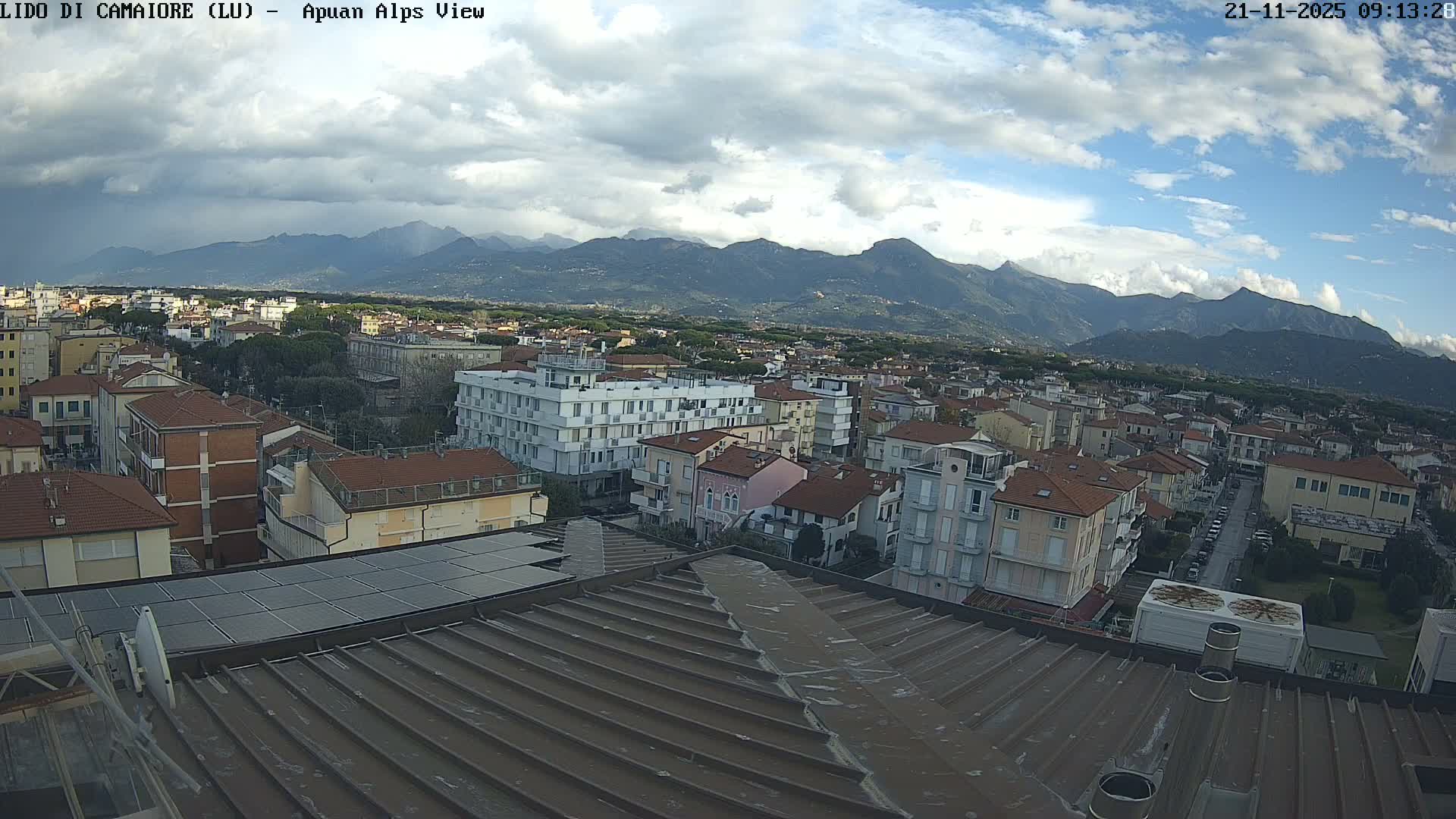 An elevated view reveals a densely built town with a mix of residential and multi-story buildings, stretching towards a distant mountain range under a dynamic, partly cloudy sky featuring both dark, heavy clouds and patches of blue.