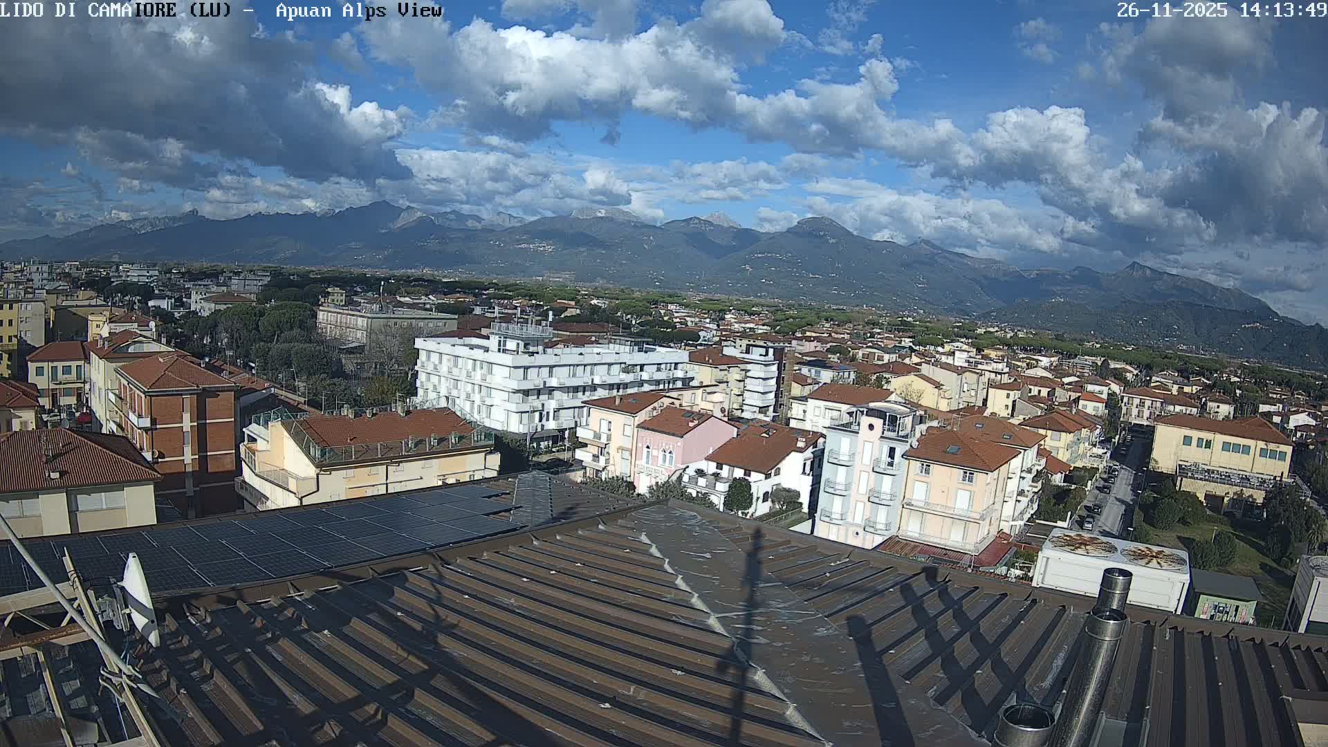 Under a sunny sky with scattered white clouds, the image presents a panoramic view of a densely built town with numerous buildings and red-tiled roofs, extending towards a grand mountain range in the distance.