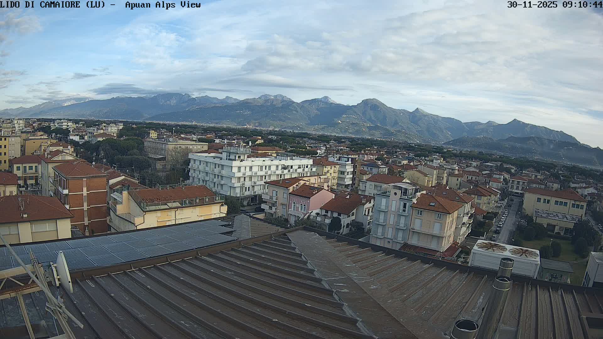 An elevated view reveals a coastal town with a mix of buildings and terracotta roofs, leading to distant rugged mountains under a partially cloudy sky, with a dark ribbed roof featuring solar panels prominent in the foreground.