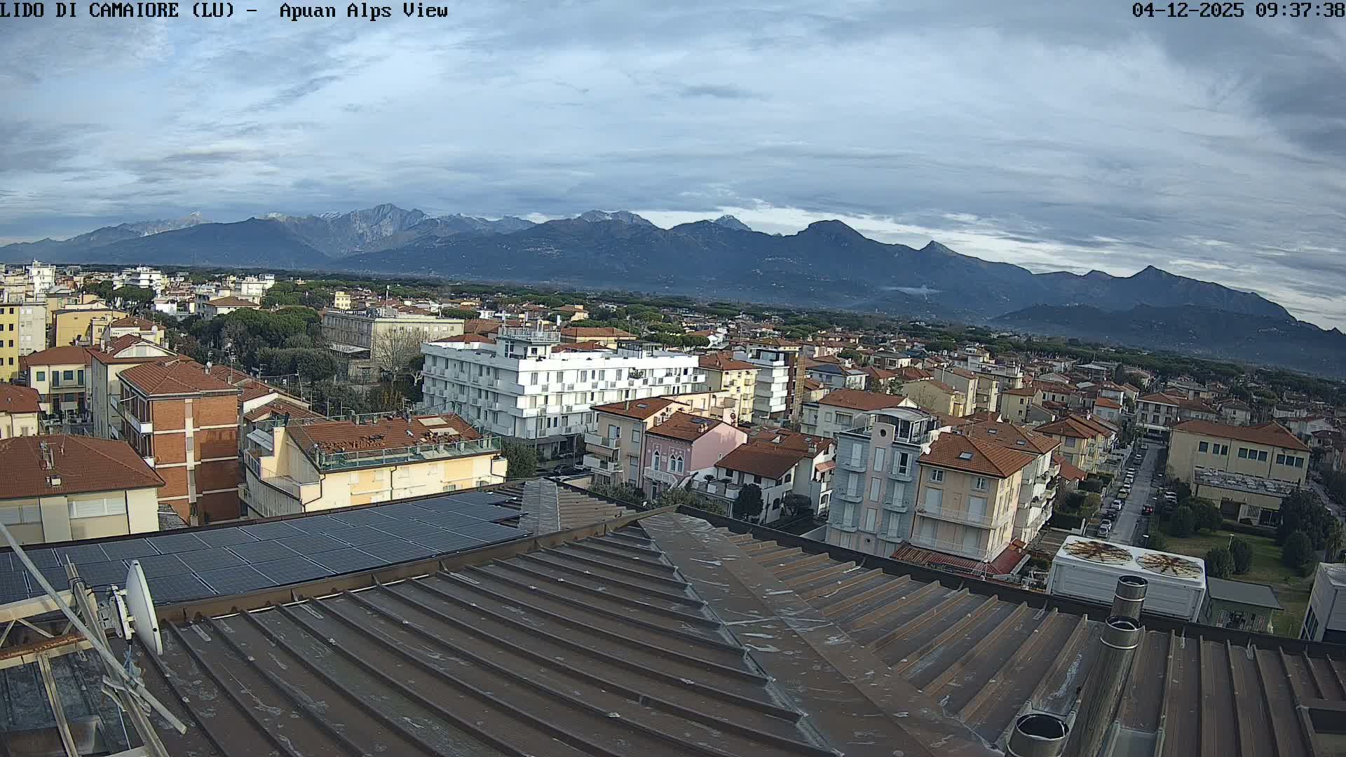 An elevated view on a cloudy day presents a townscape of buildings with predominantly terracotta roofs and scattered trees, stretching towards a distant range of partially snow-capped mountains, all observed over a metal rooftop fitted with solar panels.