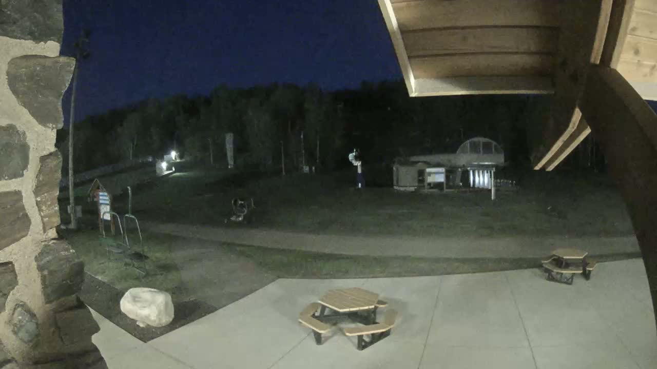 At night, a patio with two picnic tables is visible, overlooking a grassy area with some structures and trees beyond under a dark sky.