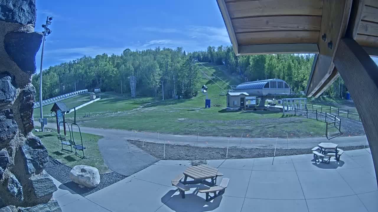 A sunny view of a ski lift and grassy hill, with picnic tables in the foreground and a stone wall to the left.