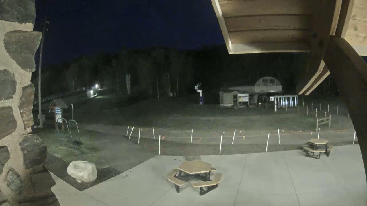 At night, a paved area with picnic tables is visible, overlooking a grassy field with a fence and some structures under a dark sky.