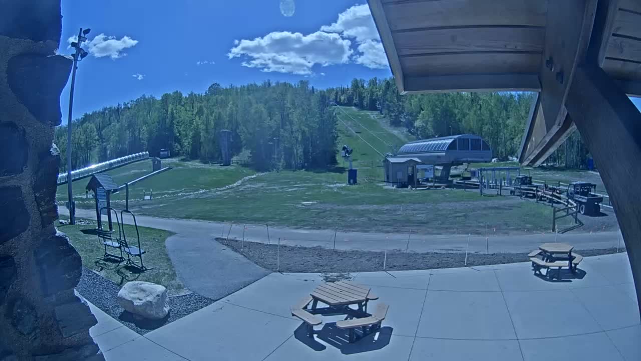 A sunny day at a ski resort shows a grassy area with a chairlift, a small building, picnic tables, and a partially visible hillside with trees in the background.