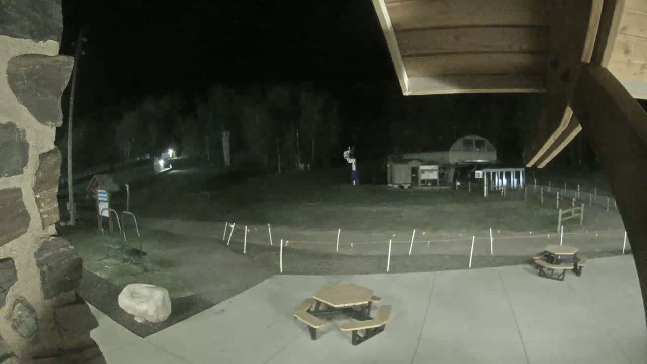 A nighttime view of a mostly empty paved area with picnic tables, a low fence, and a ski lift in the background under a dark sky.