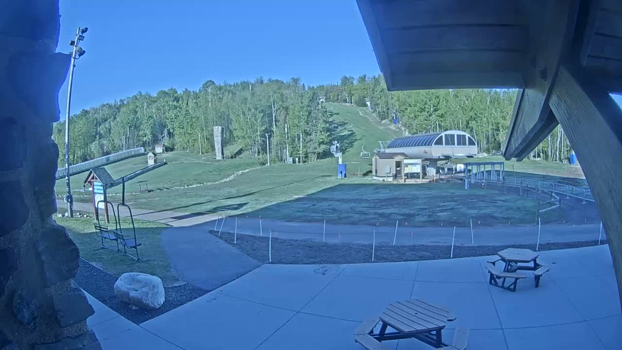 A sunny view of a ski resort's base area with a chairlift, grassy slopes, picnic tables, and a paved patio.