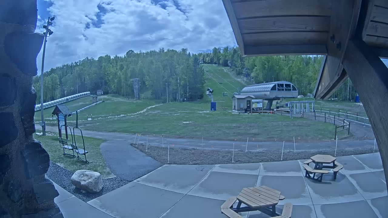 A partly cloudy day shows a ski lift and grassy area with picnic tables, a paved walkway, and a ski slope in the background.