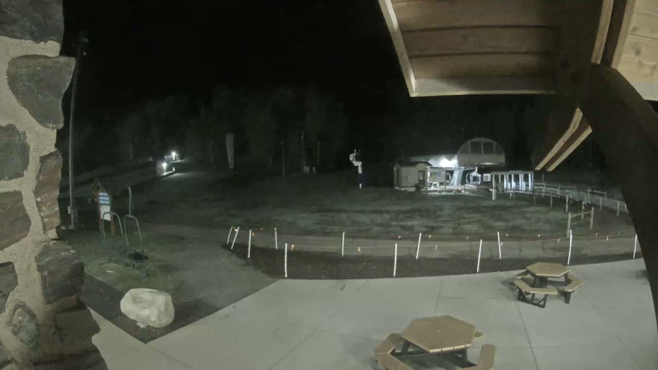 A nighttime view from under an overhang shows a snow-covered ski area with a chairlift, some small buildings, and picnic tables in a paved area.