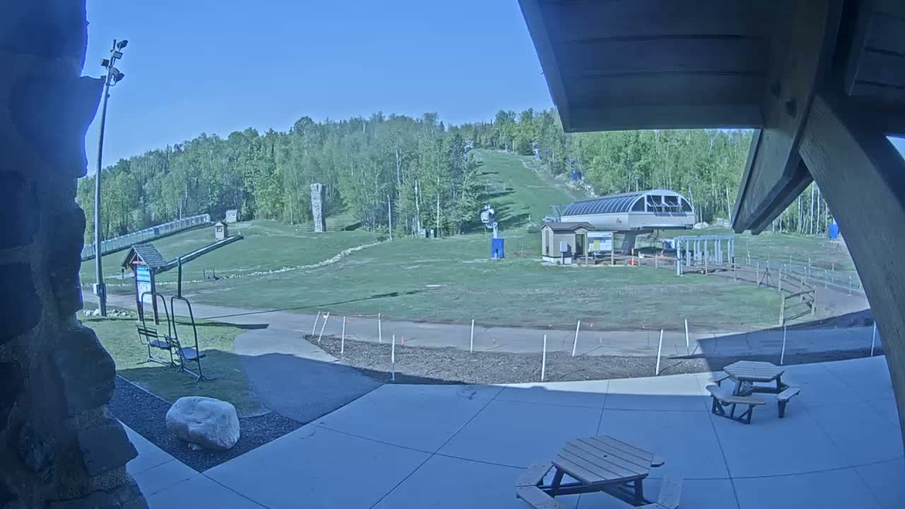 A grassy ski slope with a chairlift and picnic tables is visible on a sunny day.