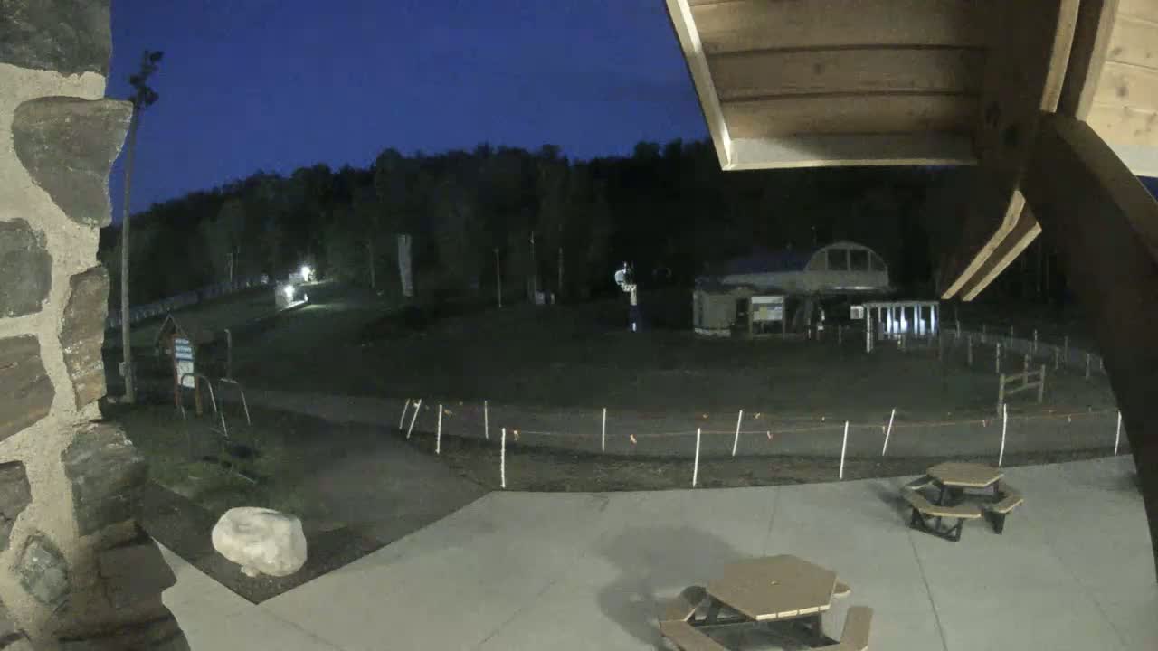 A nighttime view from a patio shows a paved area with picnic tables, a stone wall, and a grassy field beyond, under a dark blue sky.