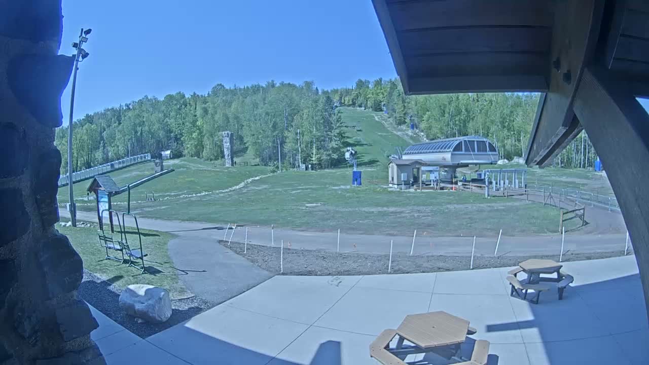 A grassy ski slope with a chairlift and a few picnic tables in the foreground on a sunny day.