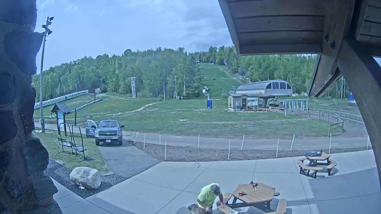 A person works on a picnic table in a paved area overlooking a grassy ski slope with a chairlift and a truck parked nearby under a partly cloudy sky.