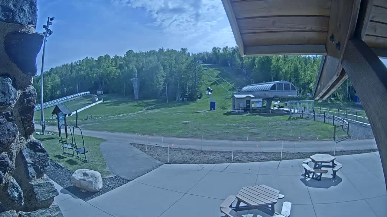 A ski lift and grassy hillside are visible under a mostly sunny sky, with picnic tables and a paved area in the foreground.