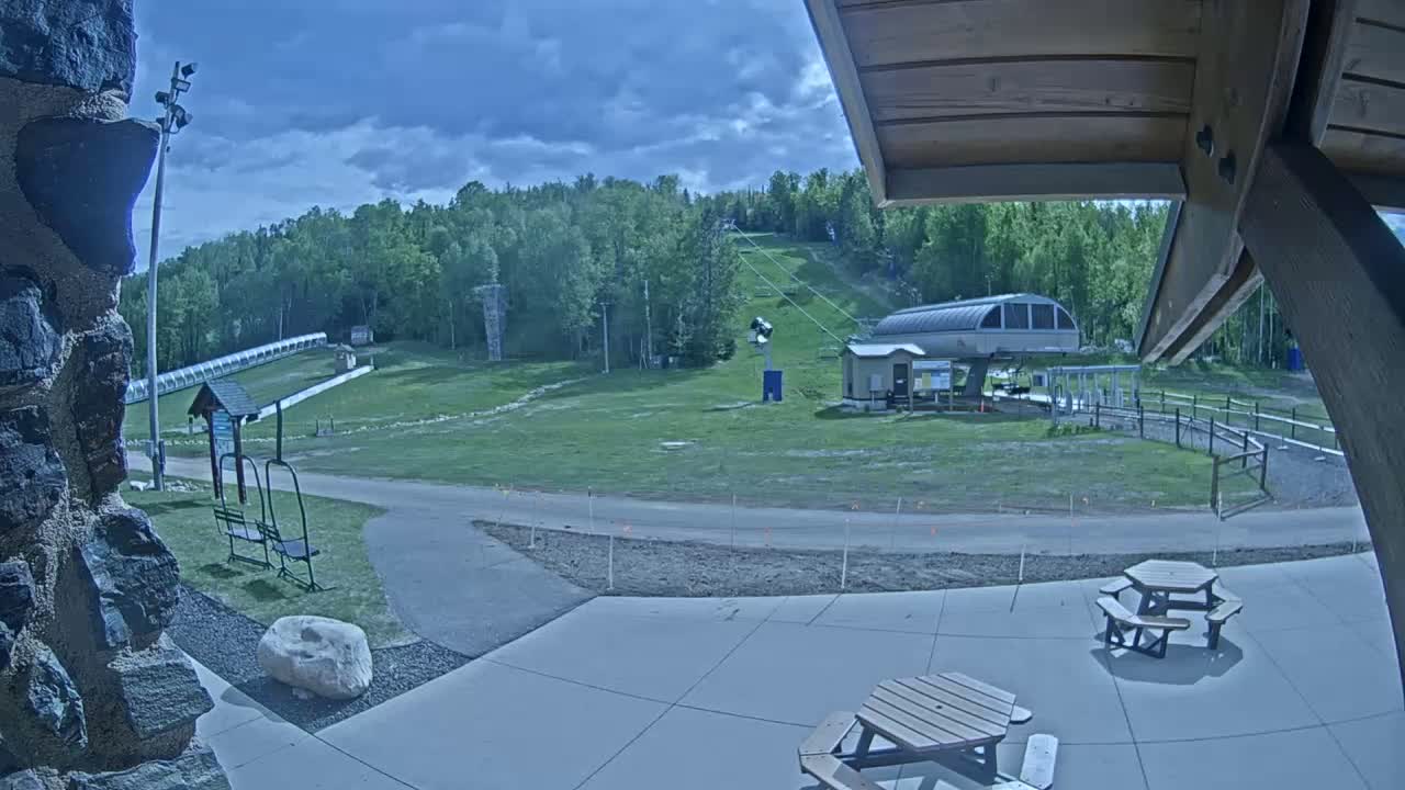 A ski lift and slope are visible beyond a paved area with picnic tables under a partly cloudy sky.