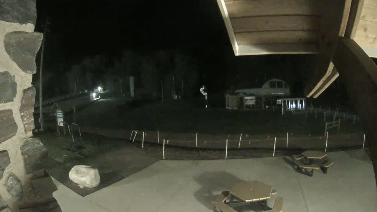 A nighttime view from under a covered structure shows a paved area with picnic tables, a stone wall, and a dimly lit grassy expanse beyond a low fence.