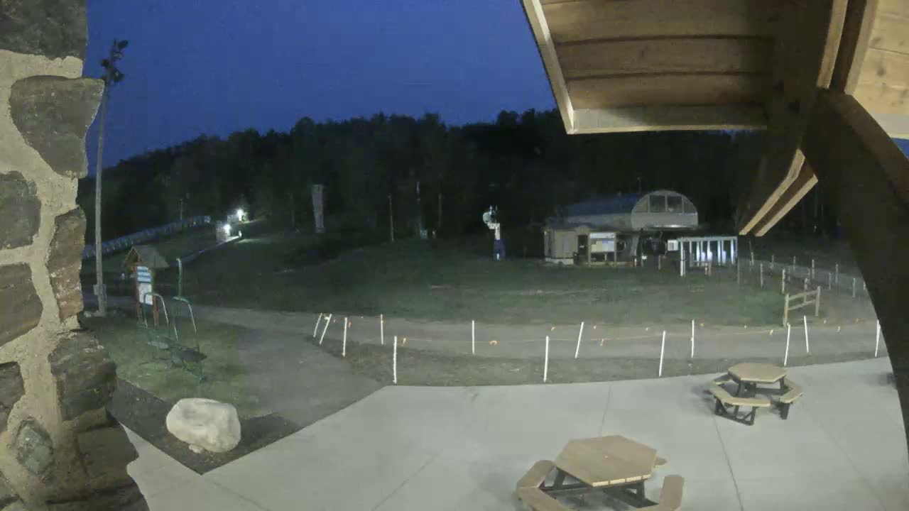 A nighttime view of an outdoor area with picnic tables, a paved path, and a building in the background under a dark blue sky.