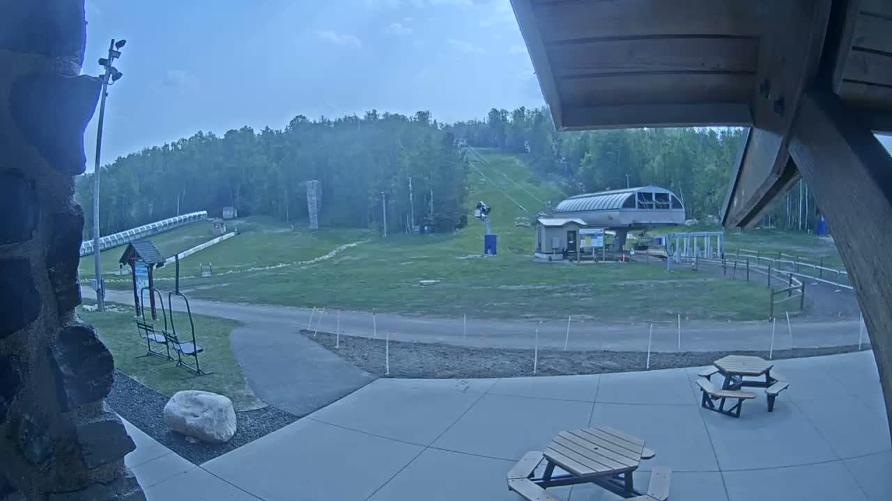 A ski lift and grassy slopes are visible under a mostly clear sky, with picnic tables and a paved area in the foreground.