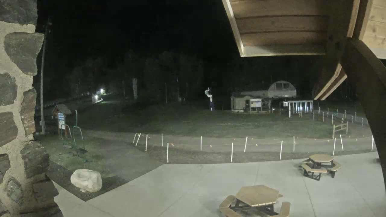 A nighttime view from a stone structure shows a paved area with picnic tables, a path winding through a grassy field towards a building and trees under a dark sky.
