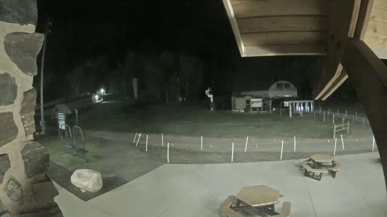 A nighttime view from under a wooden structure shows a paved area with picnic tables, a grassy field with a path and some equipment, and a building in the background under a dark sky.