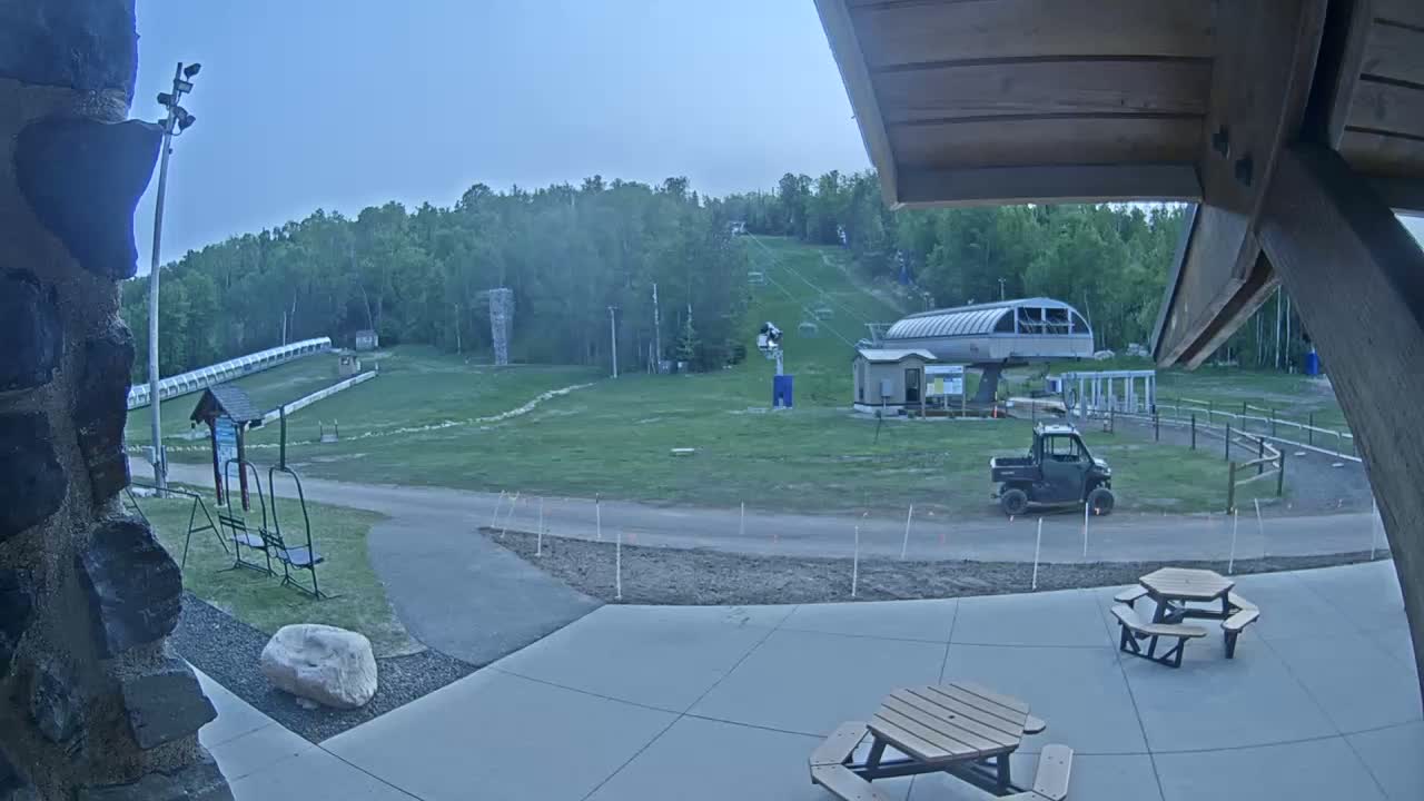 A grassy ski slope with a chairlift and a small utility vehicle is visible from a patio with picnic tables under a partly cloudy evening sky.