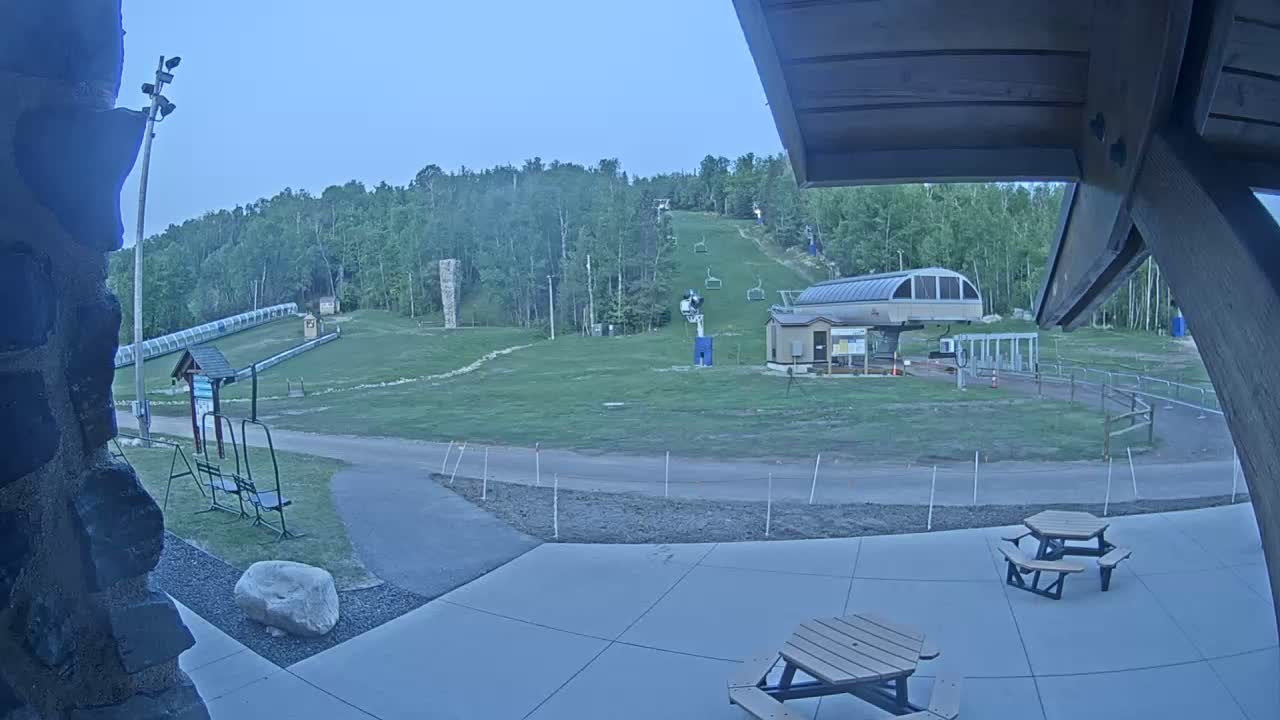 A ski resort under a twilight sky shows a grassy area with a chairlift, a small building, and picnic tables on a paved patio.