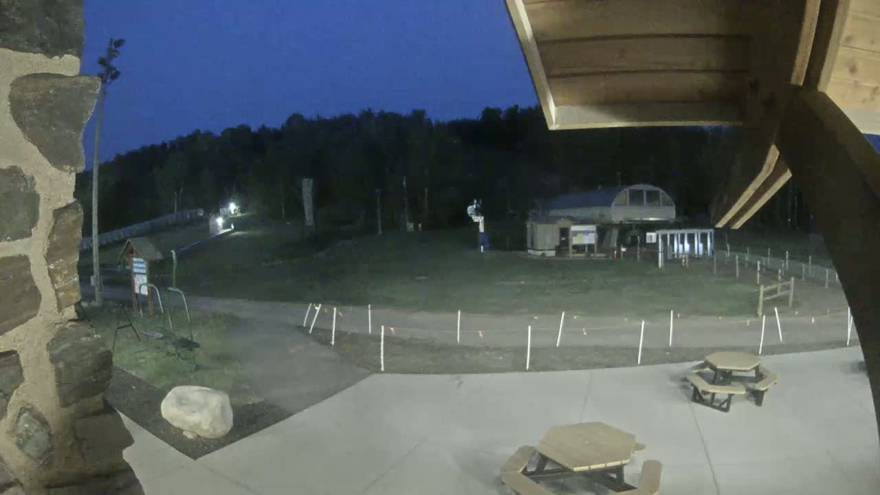 An outdoor nighttime view shows a paved area with picnic tables, a grassy field with a ski lift building in the background, and a dark forest beyond under a twilight sky.